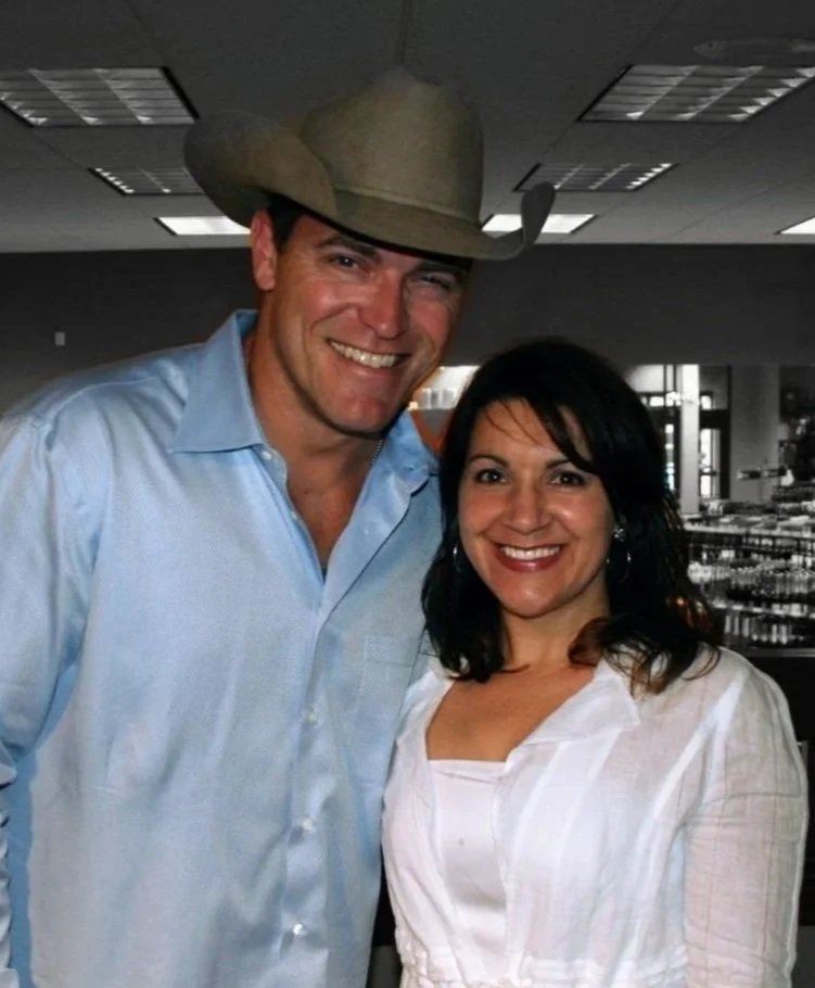 A man in a cowboy hat and a light blue shirt standing next to a woman with dark hair and a white top, both smiling indoors.