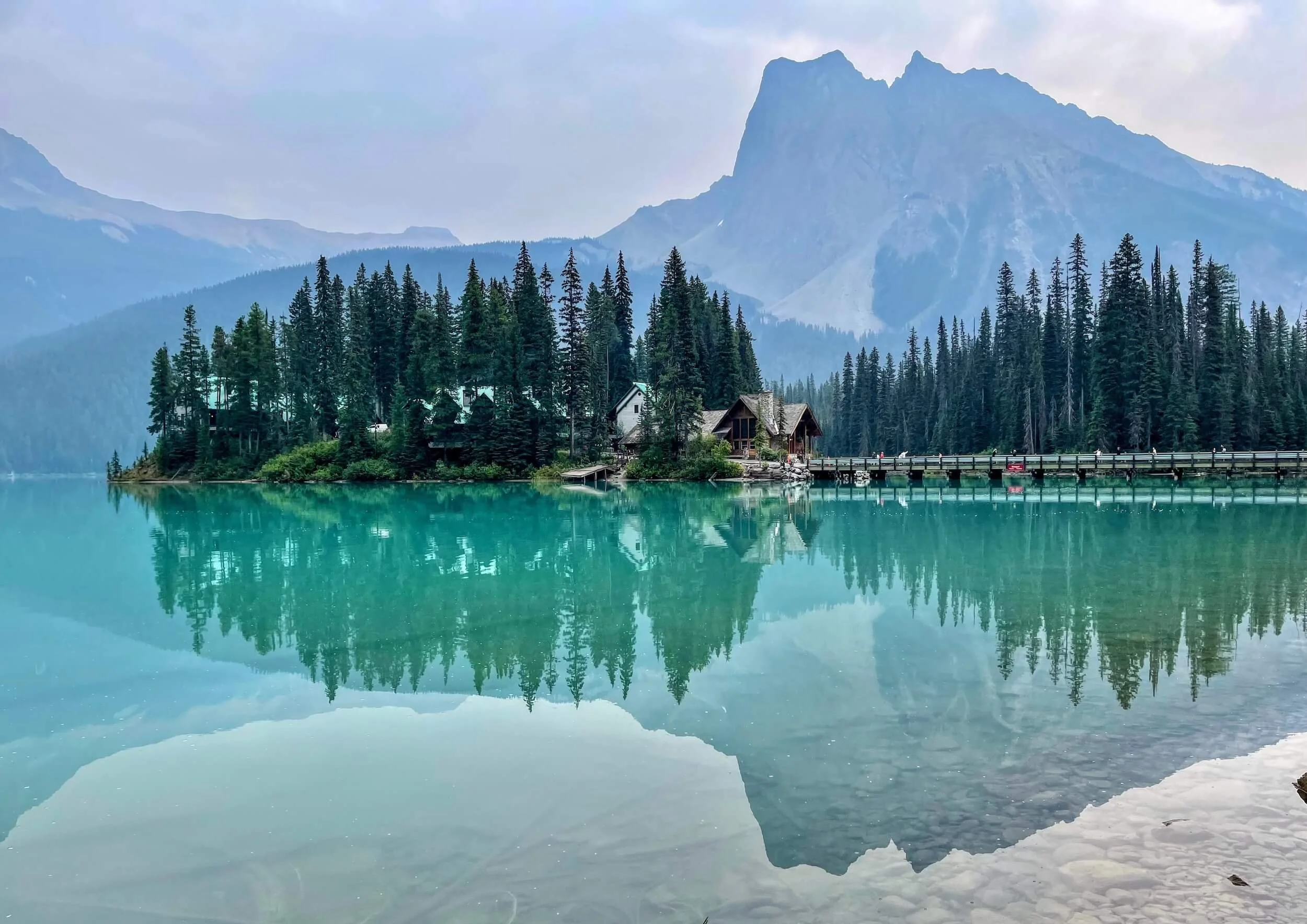A peaceful mountain lake scene with turquoise water, surrounded by dense pine trees, and tall mountains in the background under a cloudy sky.