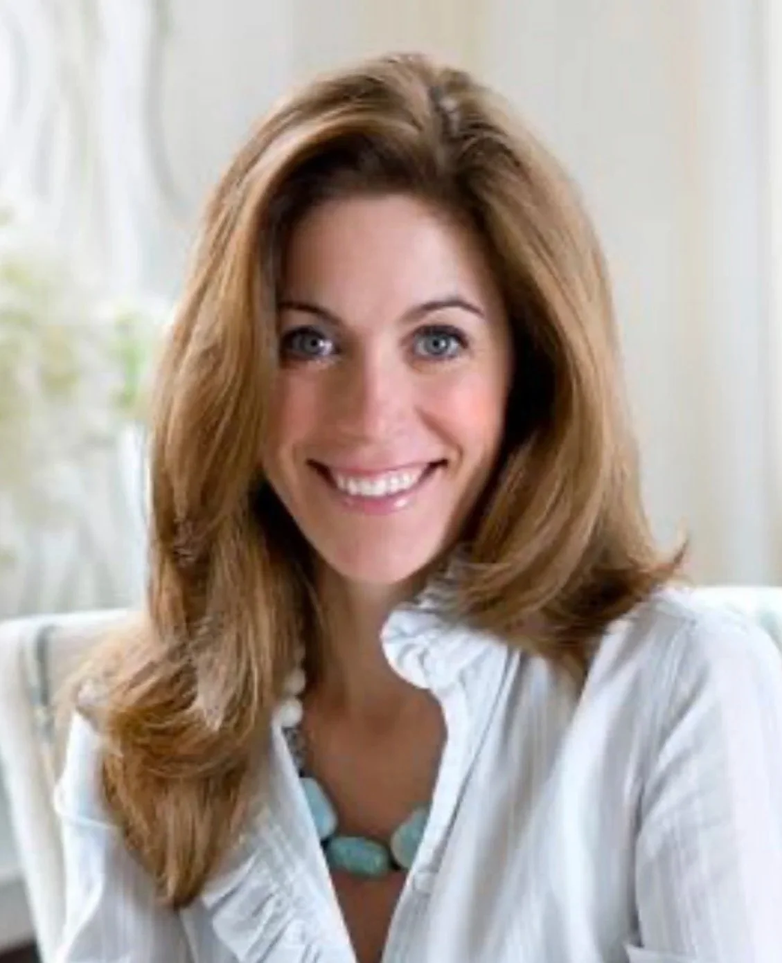A woman with shoulder-length auburn hair, blue eyes, and a bright smile, wearing a white shirt and a turquoise necklace, sitting indoors near a window.