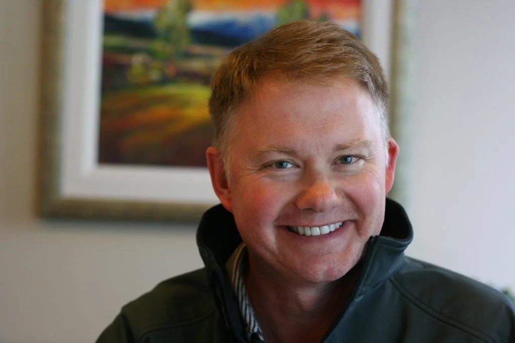 A smiling man with short, light brown hair, wearing a black jacket, indoors with a colorful landscape painting in the background.