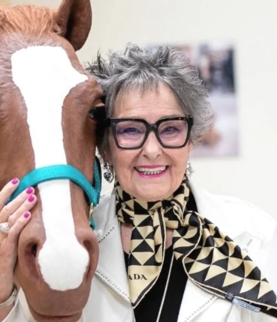 An elderly woman with short gray hair, wearing large black glasses, a cream jacket, and a black and beige geometric patterned scarf, smiling while holding a brown and white horse with her hand.