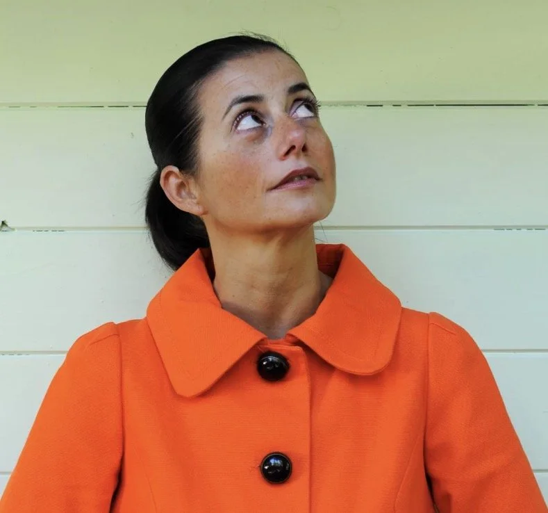 Woman with dark hair tied back, wearing an orange coat with large black buttons, looking upward with a neutral expression, standing in front of a light green wooden wall.