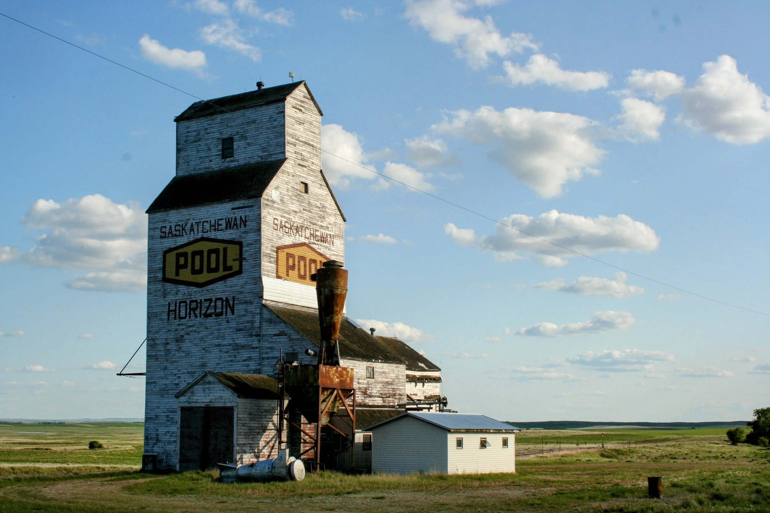 Old weathered wooden grain elevator with painted signs reading 'Saskatchewan Pool' and 'Horizon' under a partly cloudy sky, located in a rural area with flat fields and a small white building nearby.
