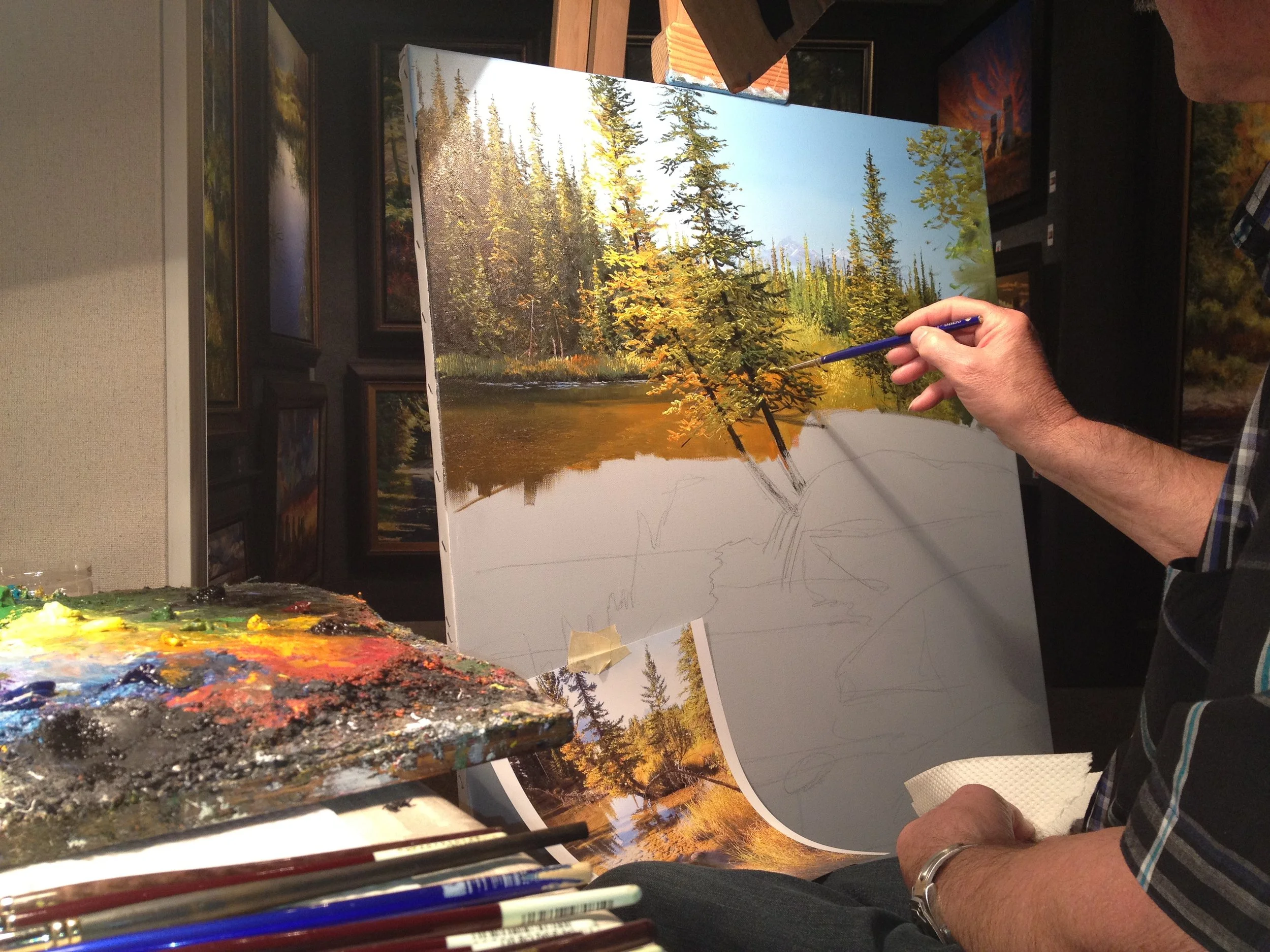 A person painting a landscape scene of trees and water on a canvas. The person is holding a paintbrush and working on the painting, which is set on an easel. The palette with various colors of paint is visible in the foreground.