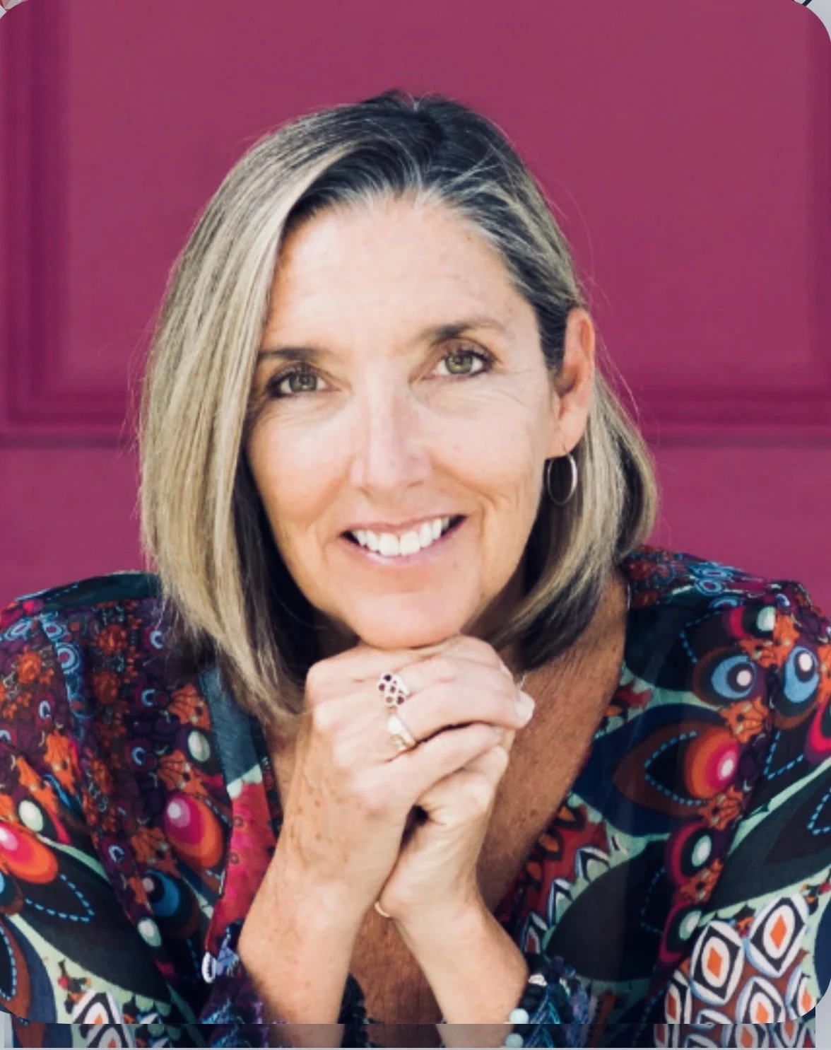 A smiling woman with short, layered blonde hair, wearing hoop earrings and a colorful patterned top, resting her chin on her hands in front of a pink background.