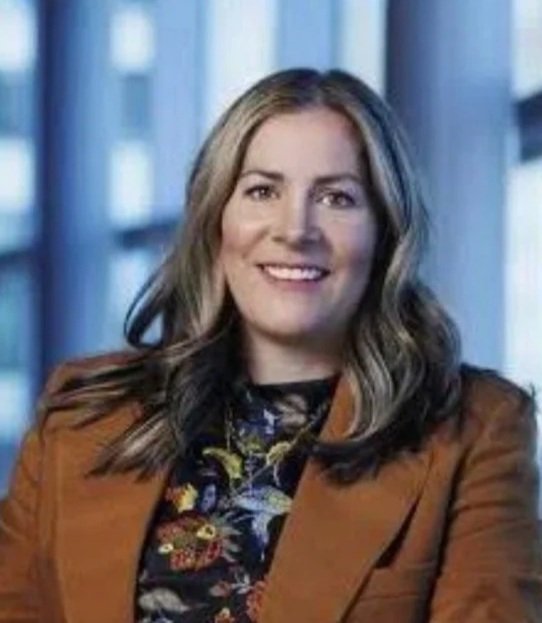 Portrait of a woman with blonde hair, wearing a floral top and brown blazer, smiling indoors.