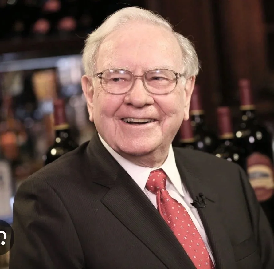 An elderly man in a black suit, white shirt, and red tie, smiling while wearing glasses, in a setting with bottles of wine in the background.