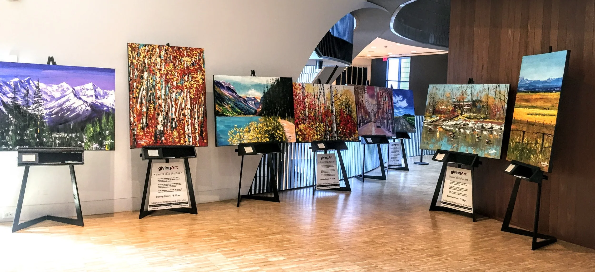An art gallery exhibition displaying eight landscape paintings on easels, featuring mountains, trees, lakes, and fields, with informational placards below each artwork.