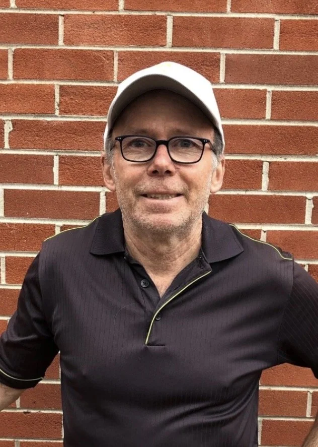 A man wearing a white cap, glasses, and a black polo shirt standing in front of a brick wall