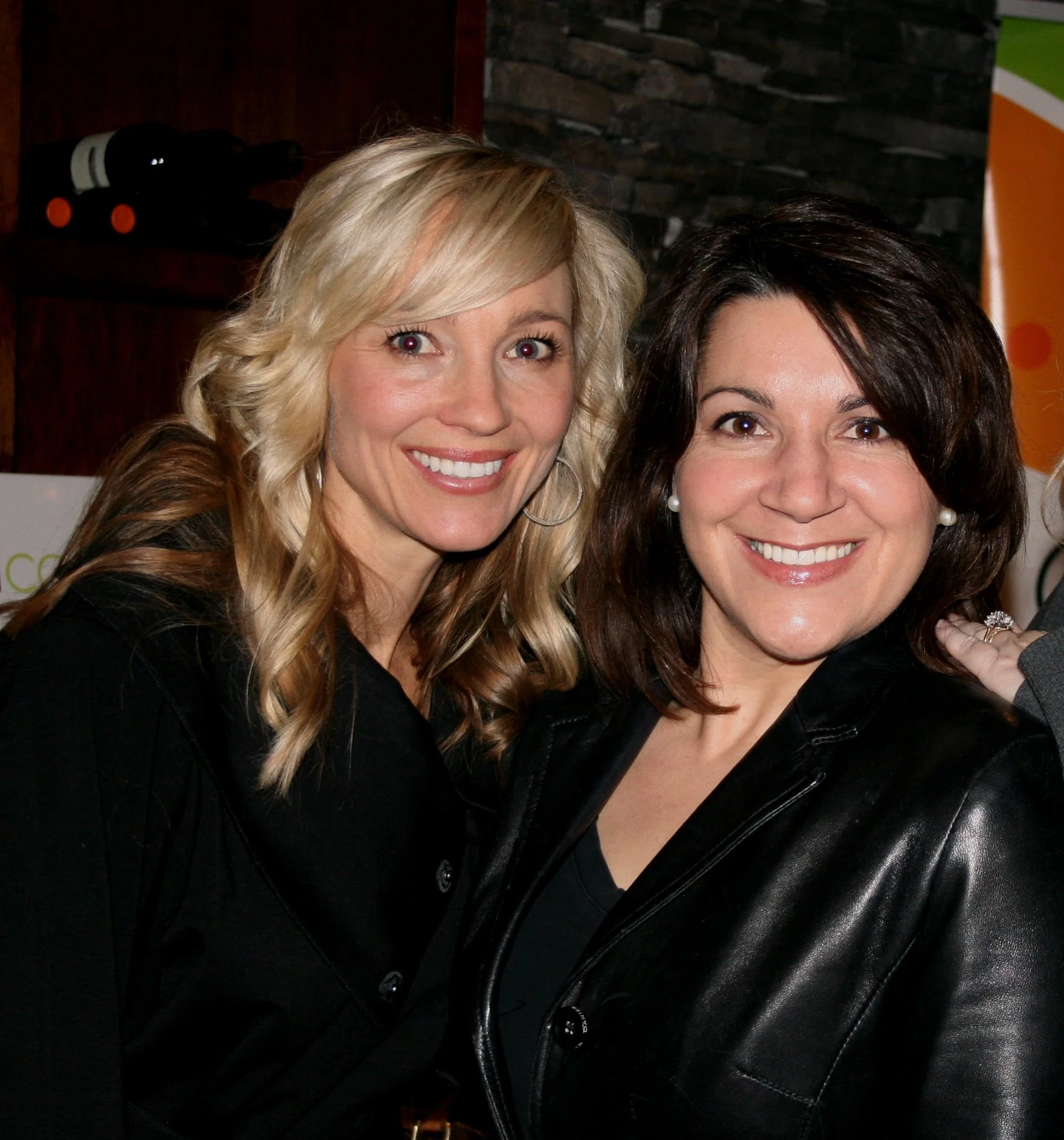 Two women smiling for a photo, one with blonde hair and the other with dark hair, wearing black clothing in an indoor setting with a dark stone wall behind them.