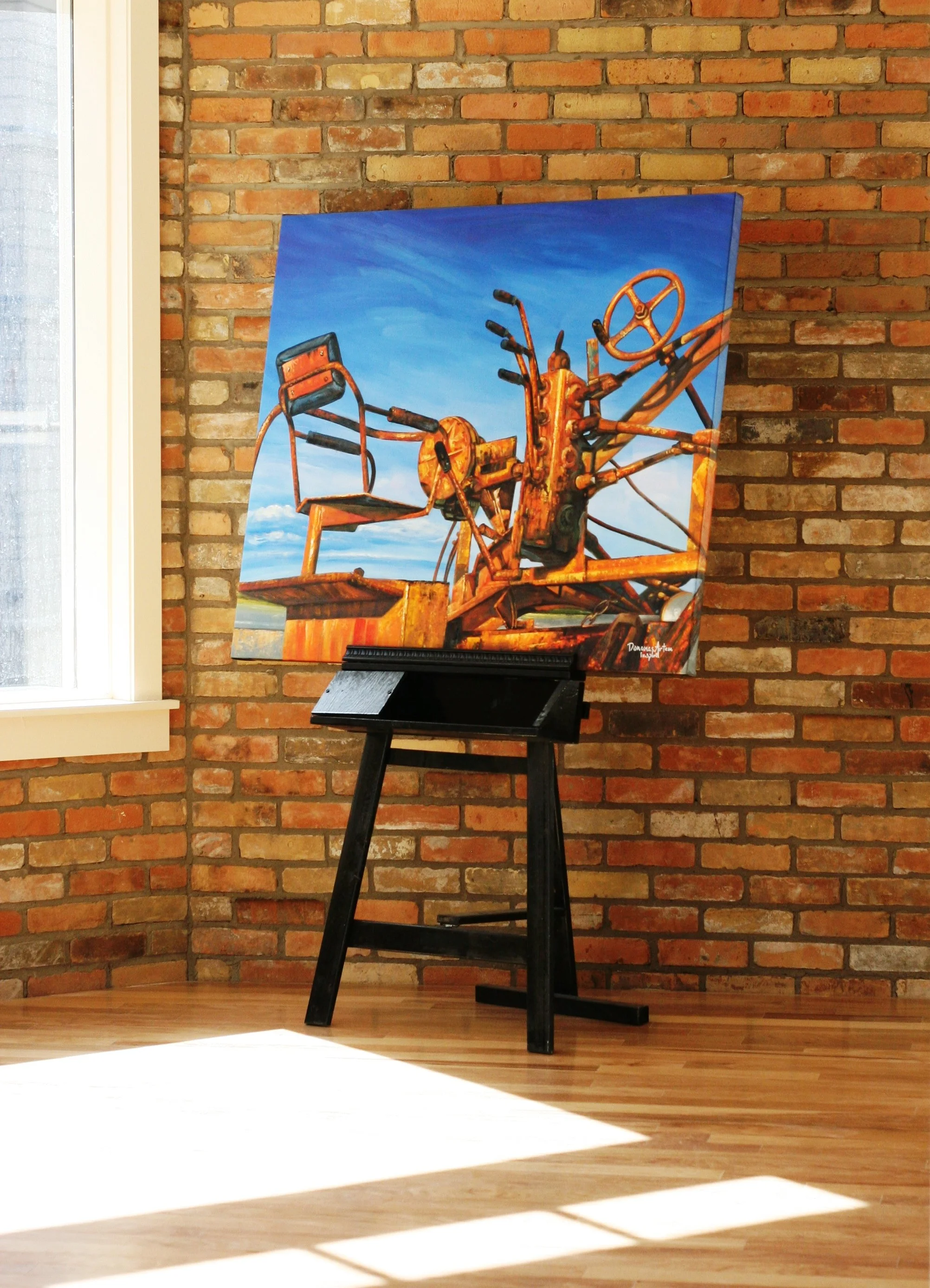 Painting of an old, rusted carnival ride with a blue sky in the background, displayed on a black easel against a brick wall with sunlight casting shadows on the wooden floor.