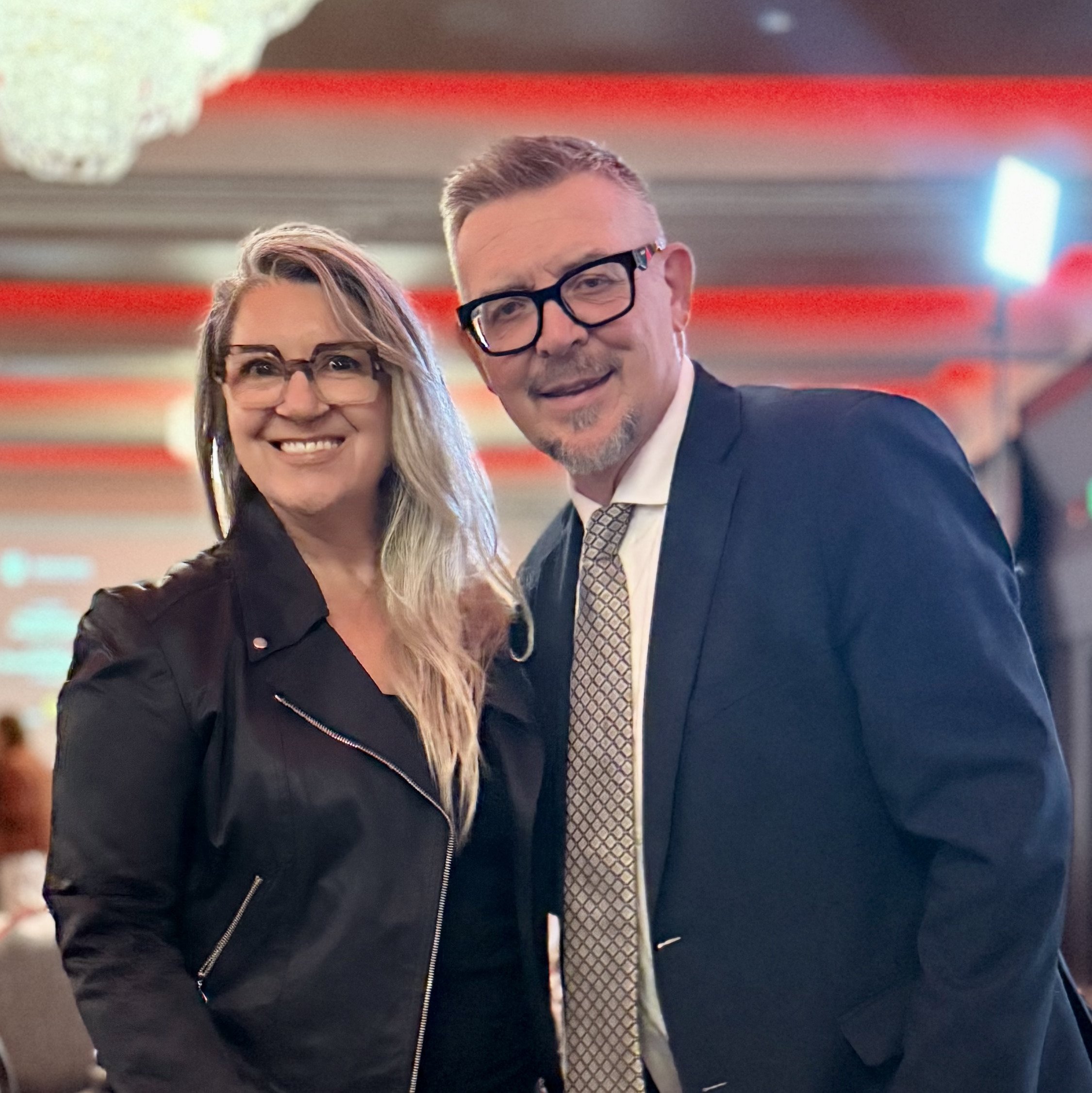 Close-up of a smiling woman with long blonde hair and glasses and a man in a suit with short hair and glasses, in an indoor setting with colorful lights.