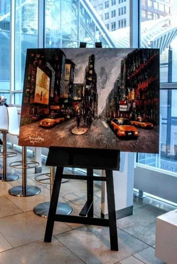 Painting of a city street scene with cars, buildings, and billboards, displayed on an easel inside a modern building with large windows.