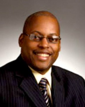 Professional headshot of a smiling man wearing glasses, a suit, and a striped tie against a plain background.