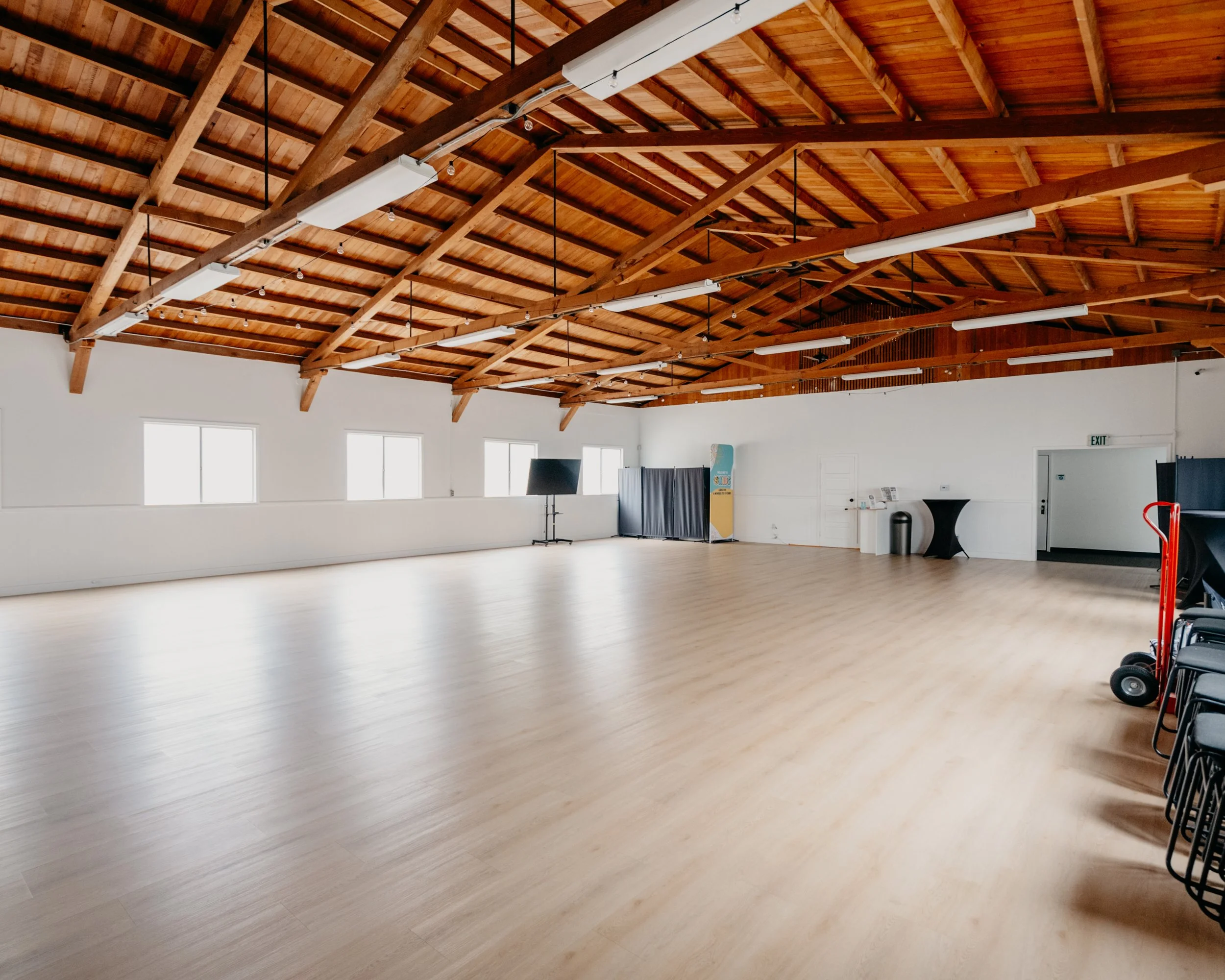 Empty meeting or event space with wooden ceiling, white walls, black chairs along the wall, and a few windows. Some equipment and a sign are visible in the room.