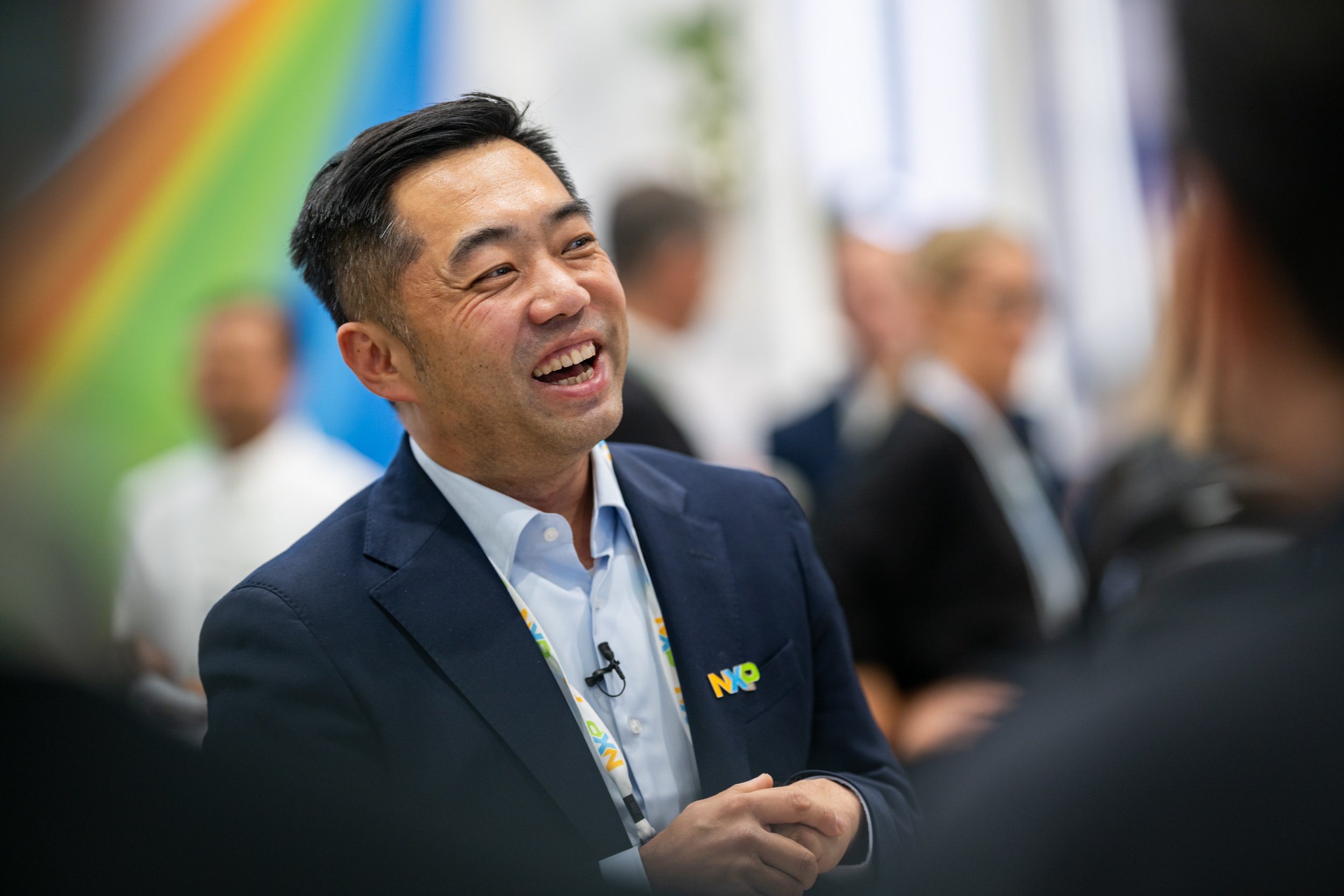 A man in a blue blazer and white shirt, smiling and talking at an indoor event with other people blurred in the background. He is wearing an event badge and a lapel pin, and appears to be engaged in conversation.
