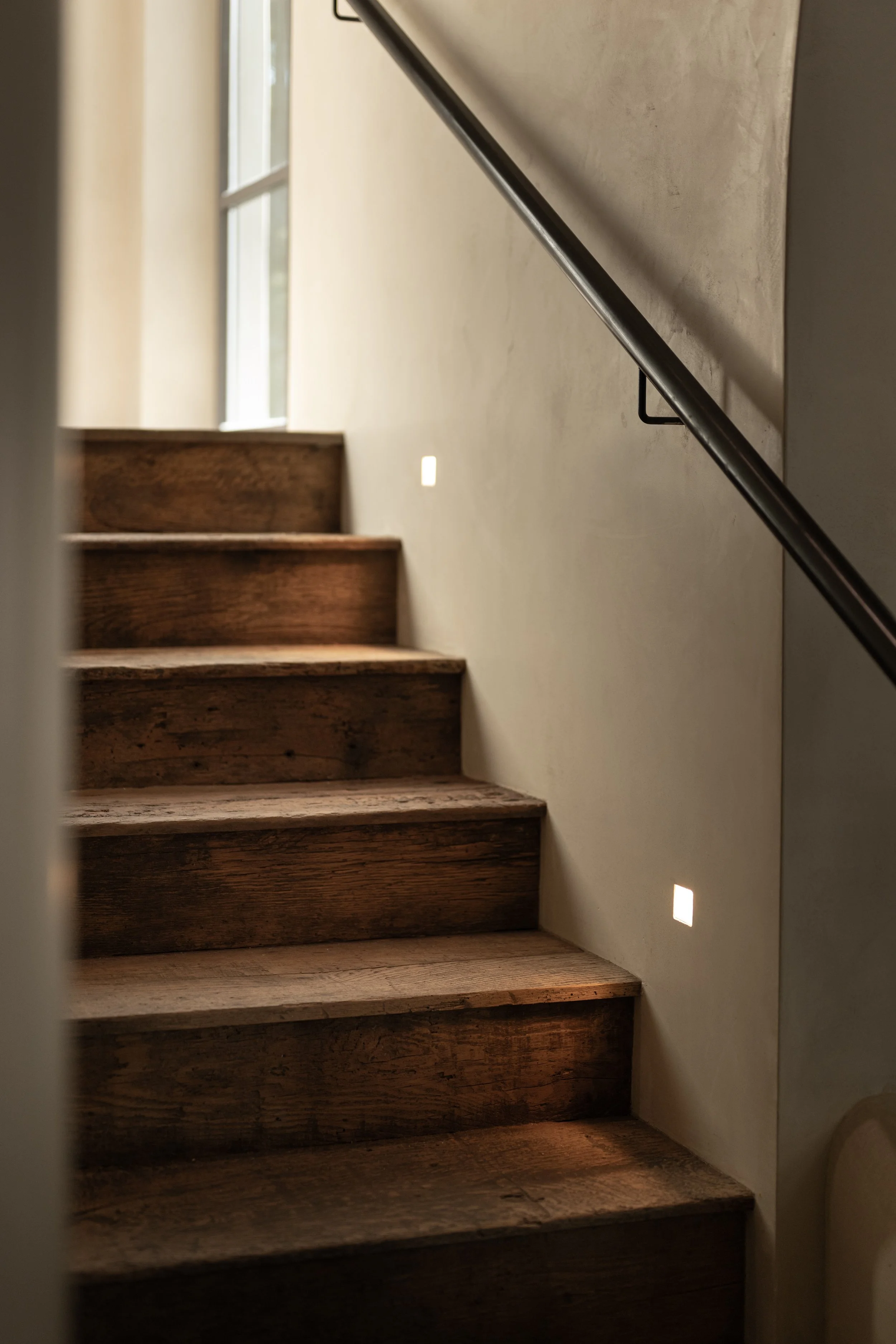 Wooden staircase with a black metal handrail next to a cream-colored wall, with small square lights illuminating the stairs from the wall.