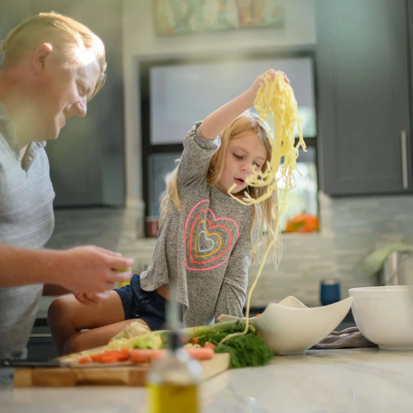 A man and a young girl in a kitchen preparing food, with the girl holding cooked pasta above her head and looking at it, while the man is standing nearby.