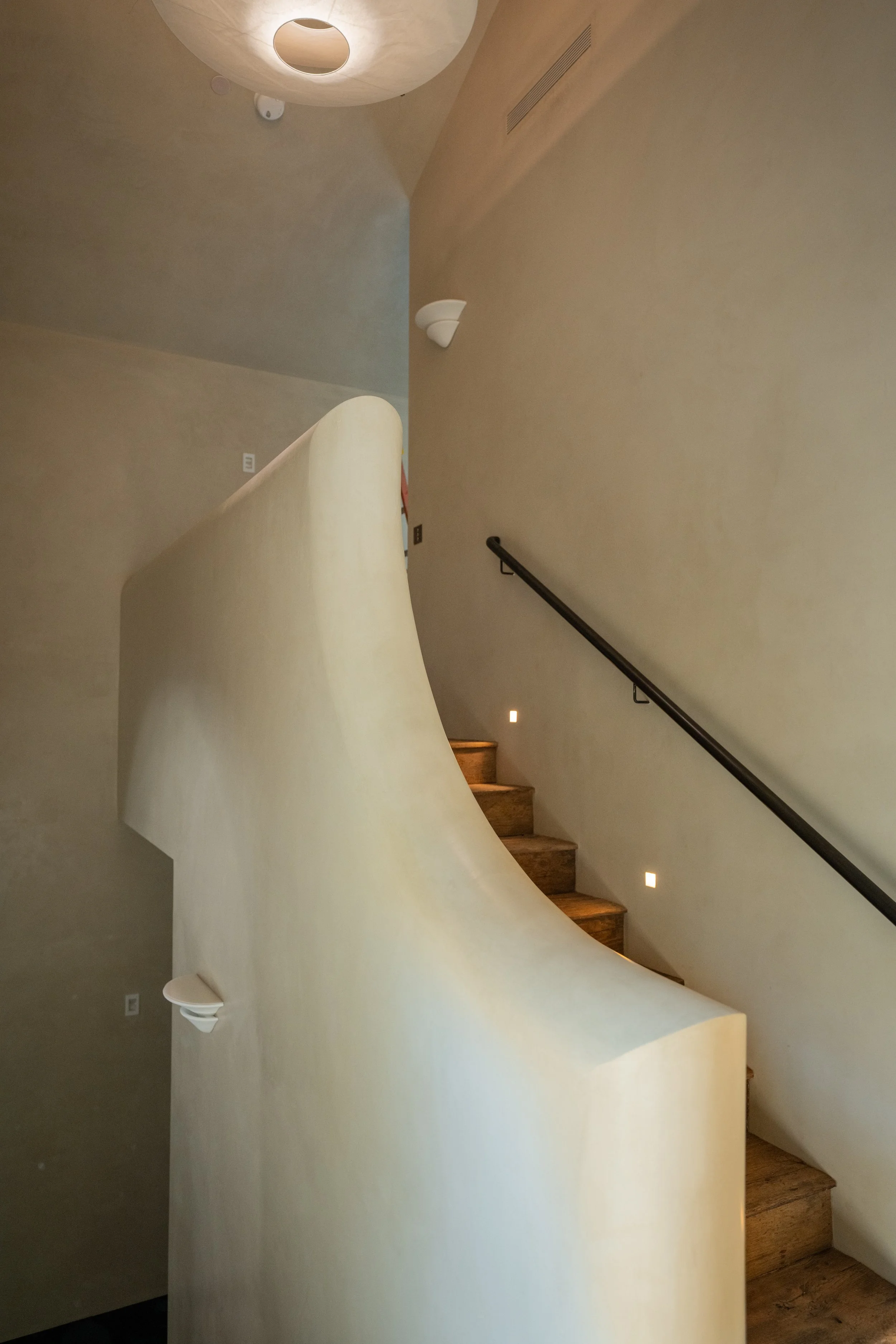 Interior view of a modern staircase with wooden steps, a black handrail, beige curved wall, and minimal lighting. The photo is taken from the bottom of the stairs looking upward.