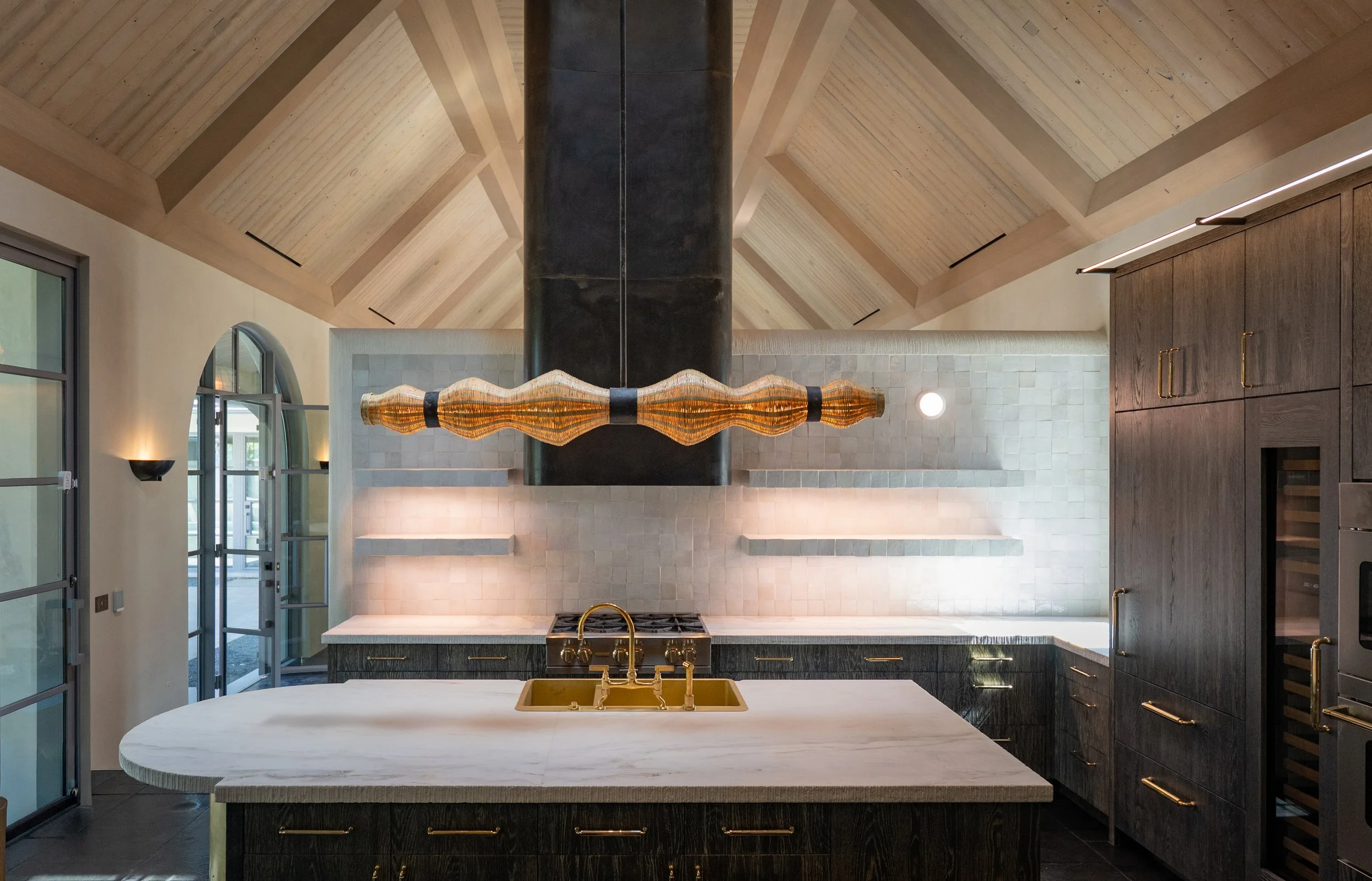 Modern kitchen with a large white island, a gold sink, dark cabinetry, wooden ceiling beams, pendant lighting, and arched windows.