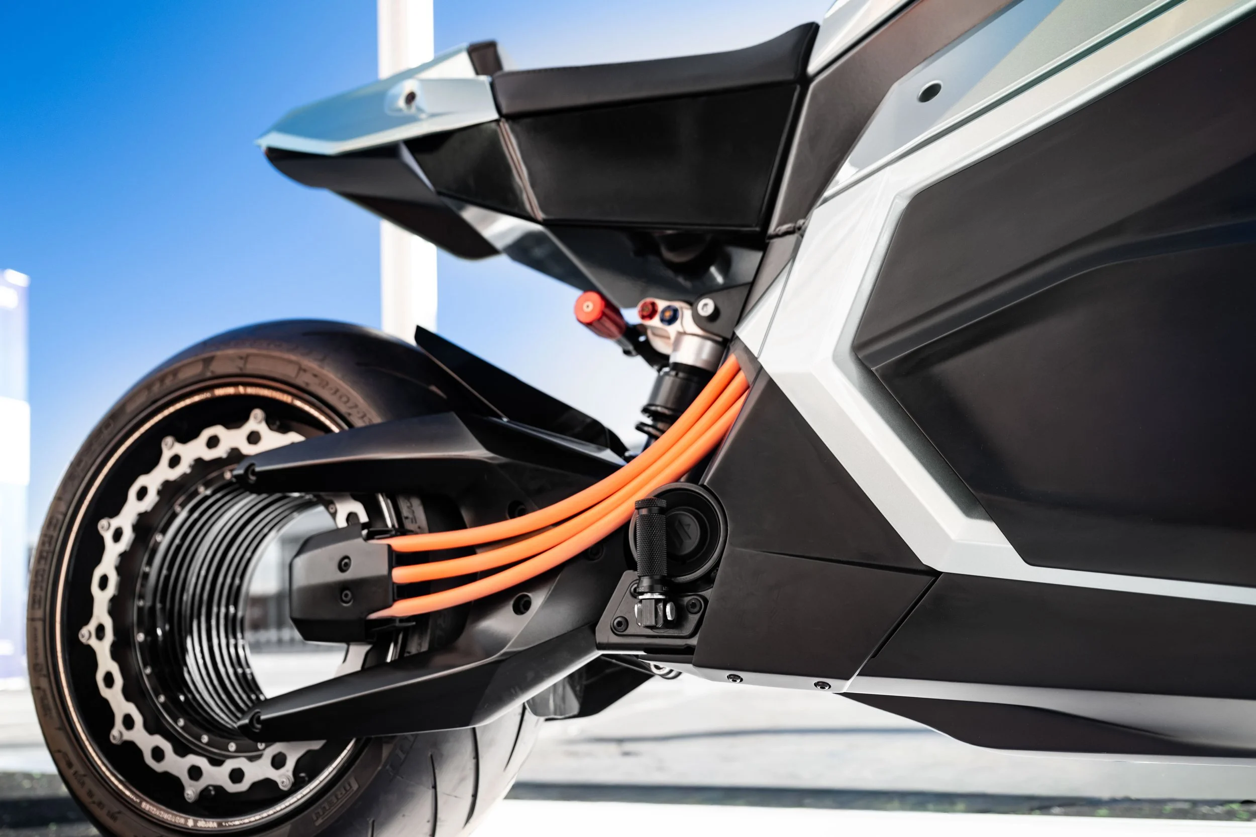 Close-up of the rear part of an electric motorcycle, showing orange cables, a black wheel, and disc brake against a blue sky background.