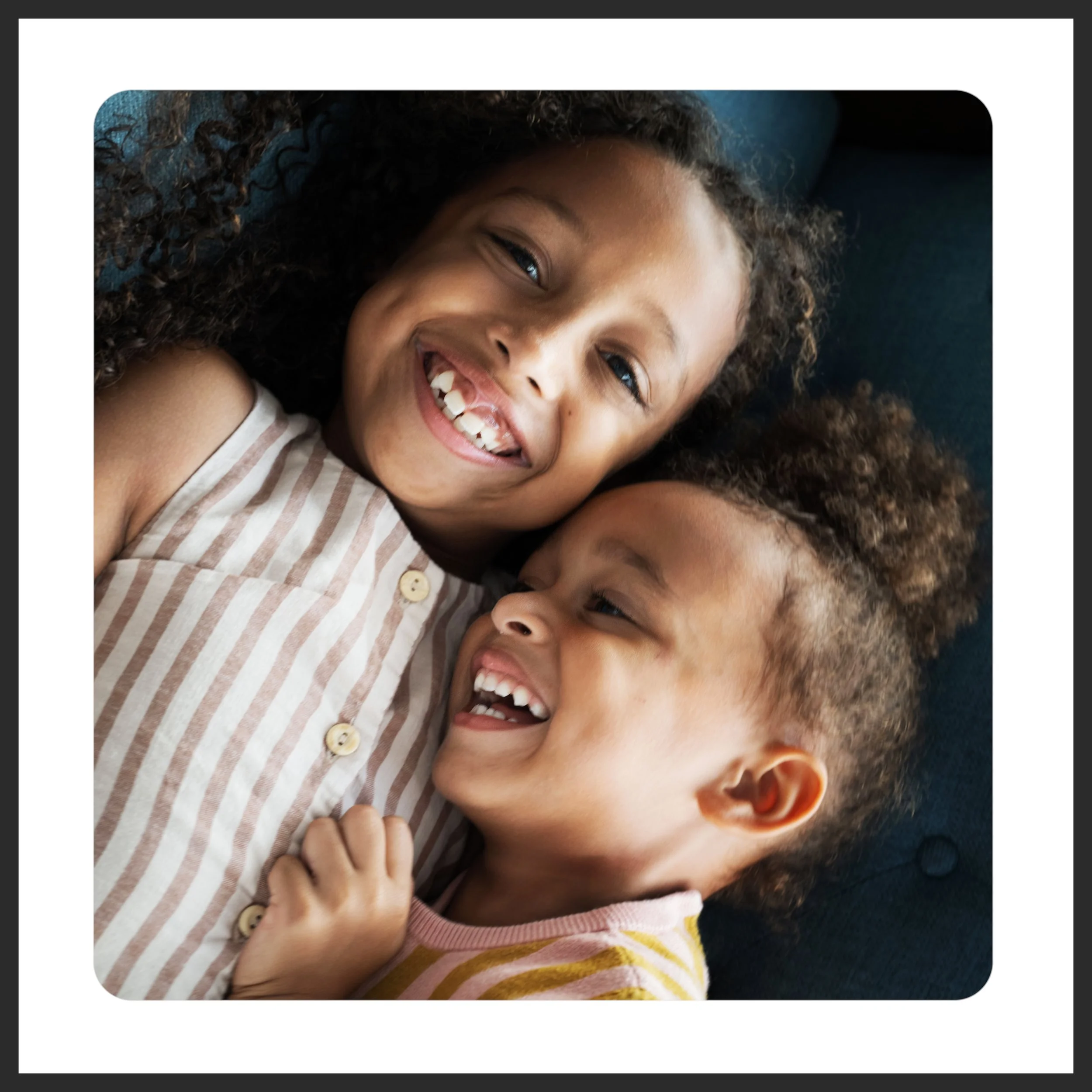 Close-up of two smiling children lying on a dark blue surface. Both children have curly hair and are sharing a joyful moment.