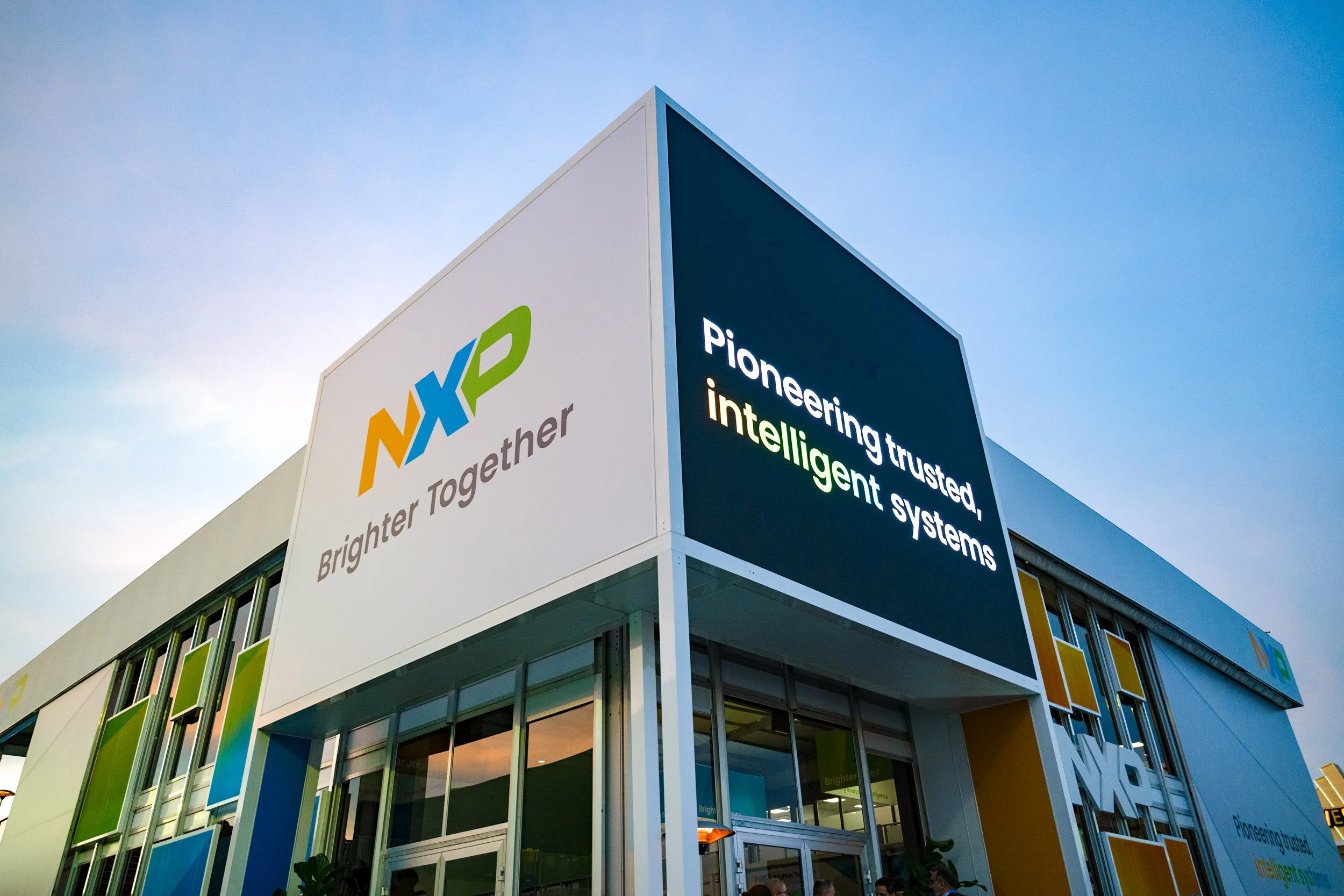 A modern building with large glass windows and colorful banners on the facade, featuring the NX Pioneering System logo and slogan about trusted, intelligent systems, with a clear sky in the background.