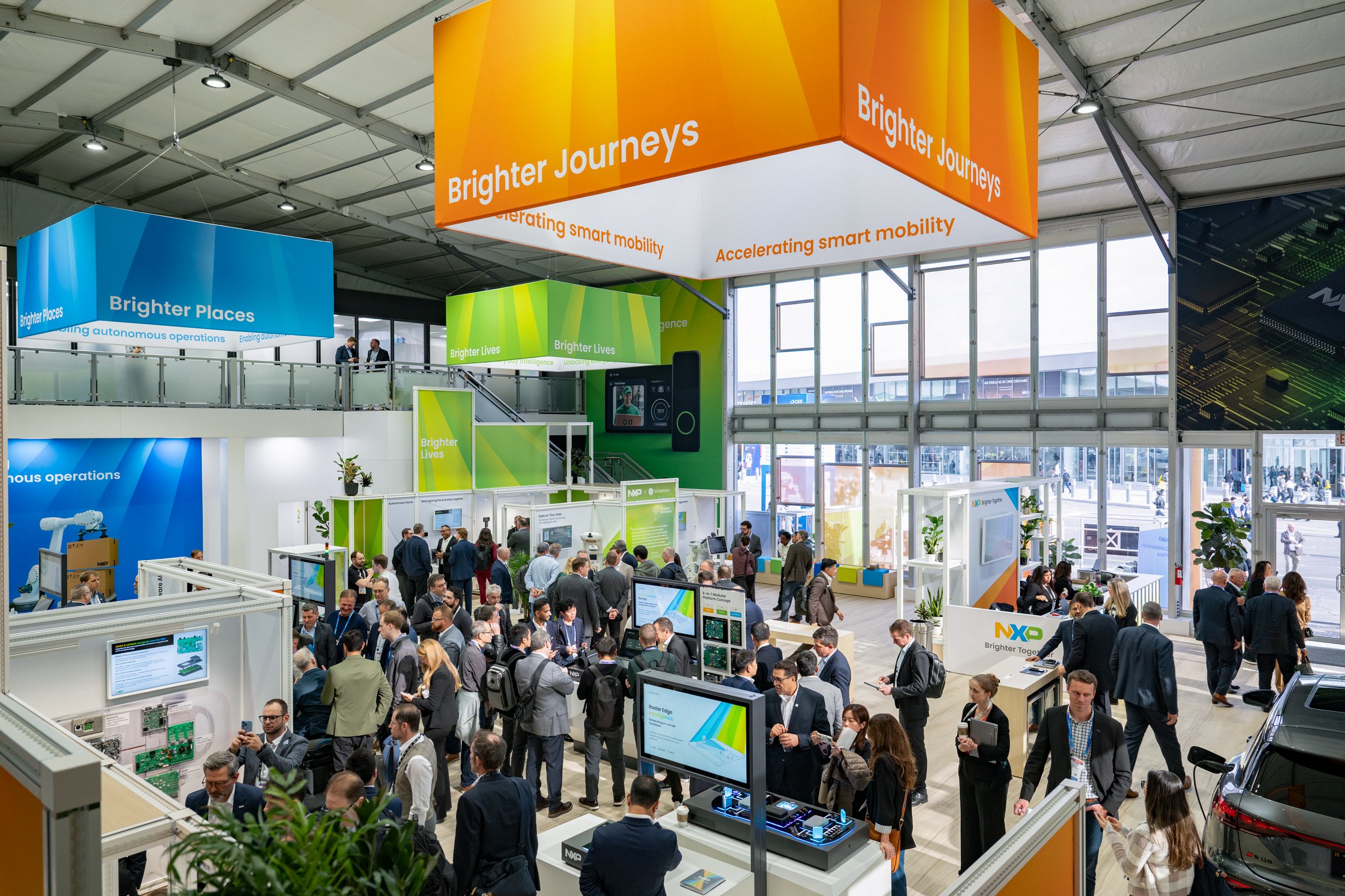 Busy conference or trade show with multiple booths and banners, attendees walking and interacting, large windows, and digital displays.