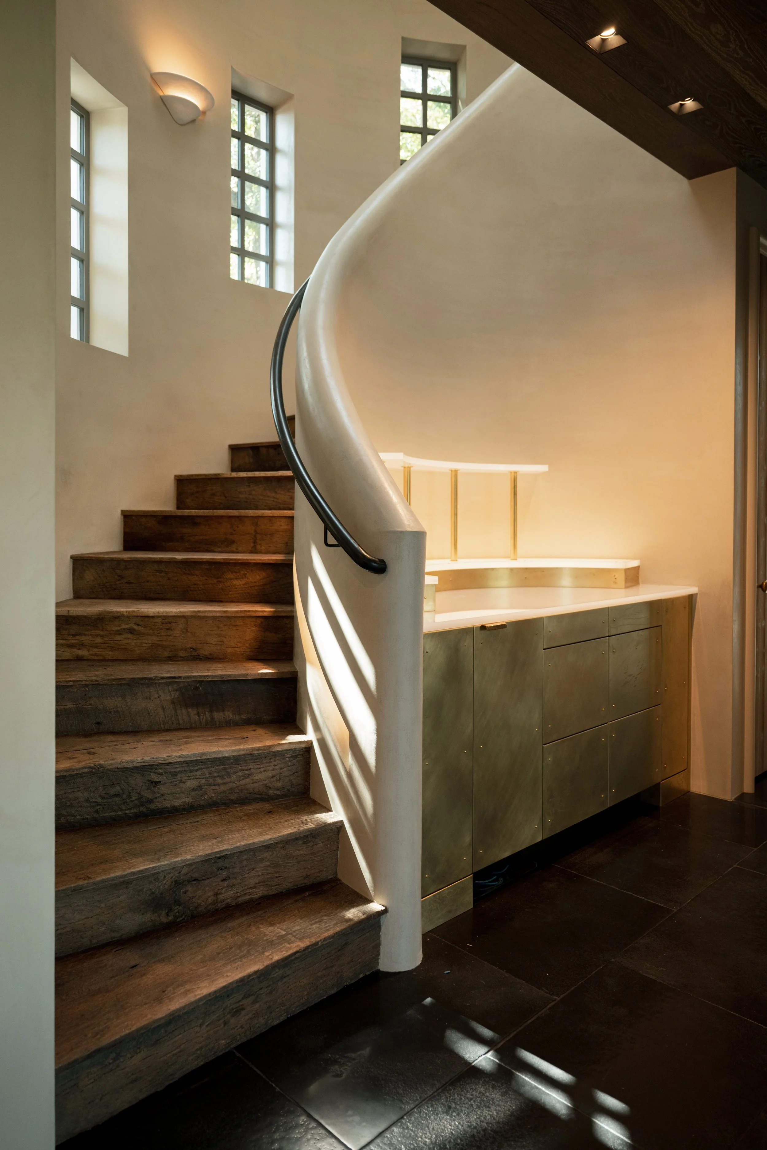 Interior view of a modern staircase with dark wooden steps, a curved white wall, and patterned black flooring, illuminated by natural light from tall, narrow windows.