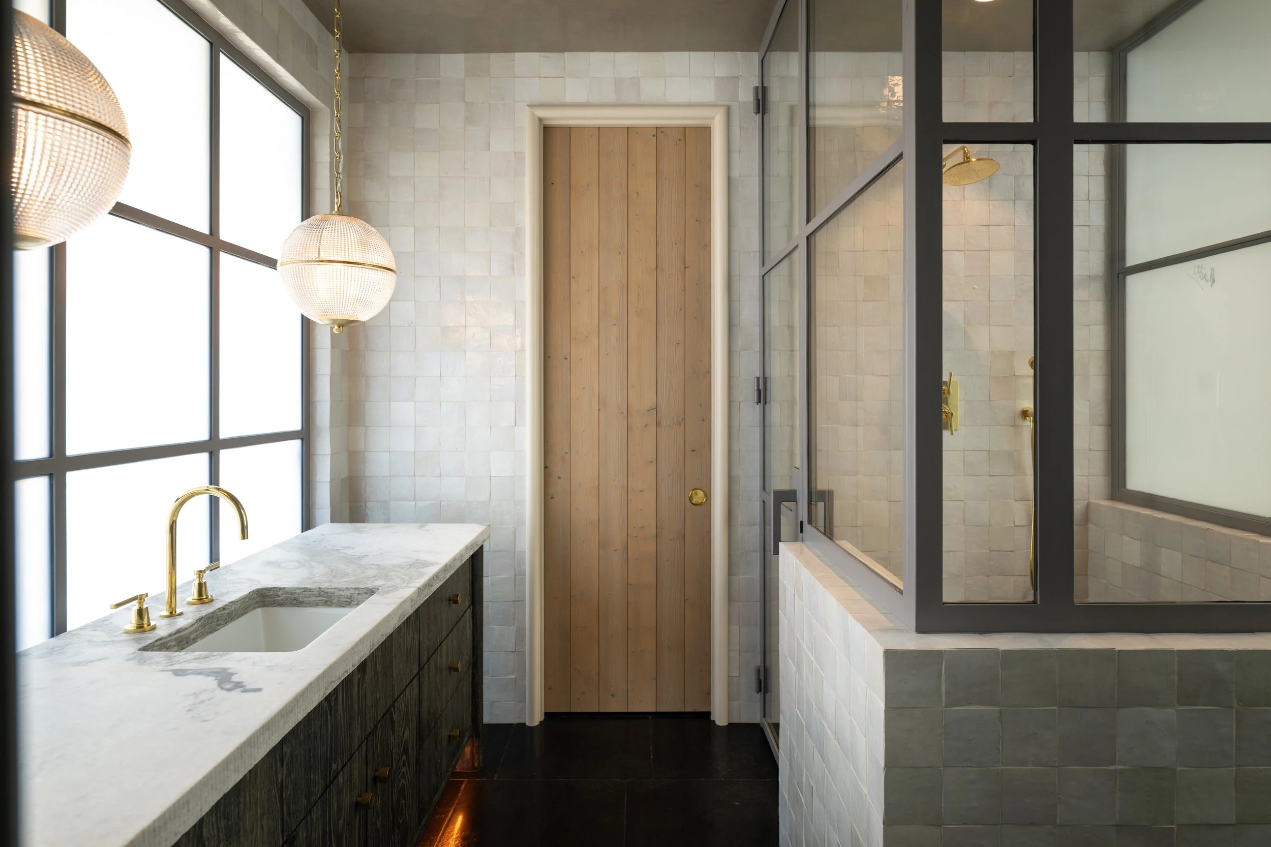 Modern bathroom with a marble countertop, gold fixtures, tiled walls, a wooden door, a glass shower enclosure, and large windows letting in natural light.