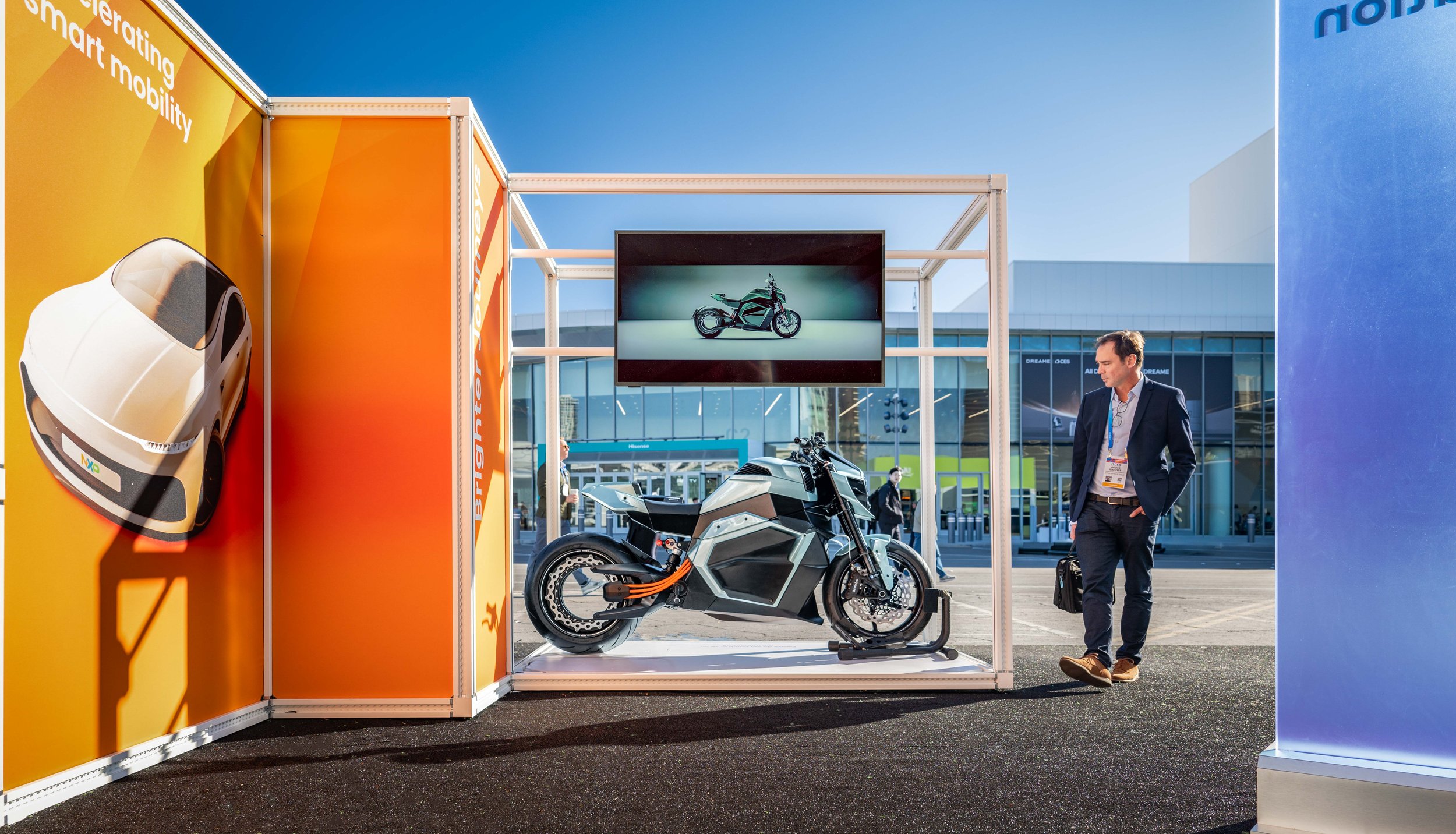 A man in a suit stands next to a futuristic black and gray electric motorcycle displayed on a stand at an outdoor event. Behind him, a large screen shows an image of a similar motorcycle. To the left, colorful panels and a white, closed car display a