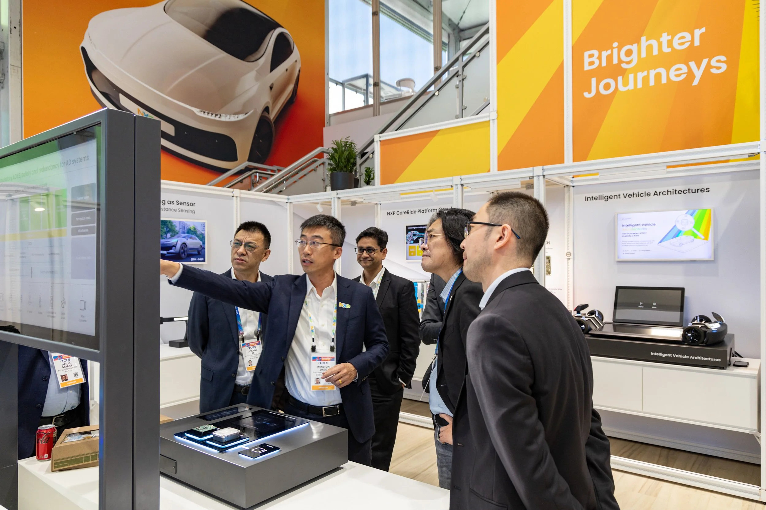 A group of men in formal attire are gathered around a display table at an exhibition, with one man explaining something on a screen. The background features large orange and yellow banners with the words 'Brighter Journeys' and images of vehicles and