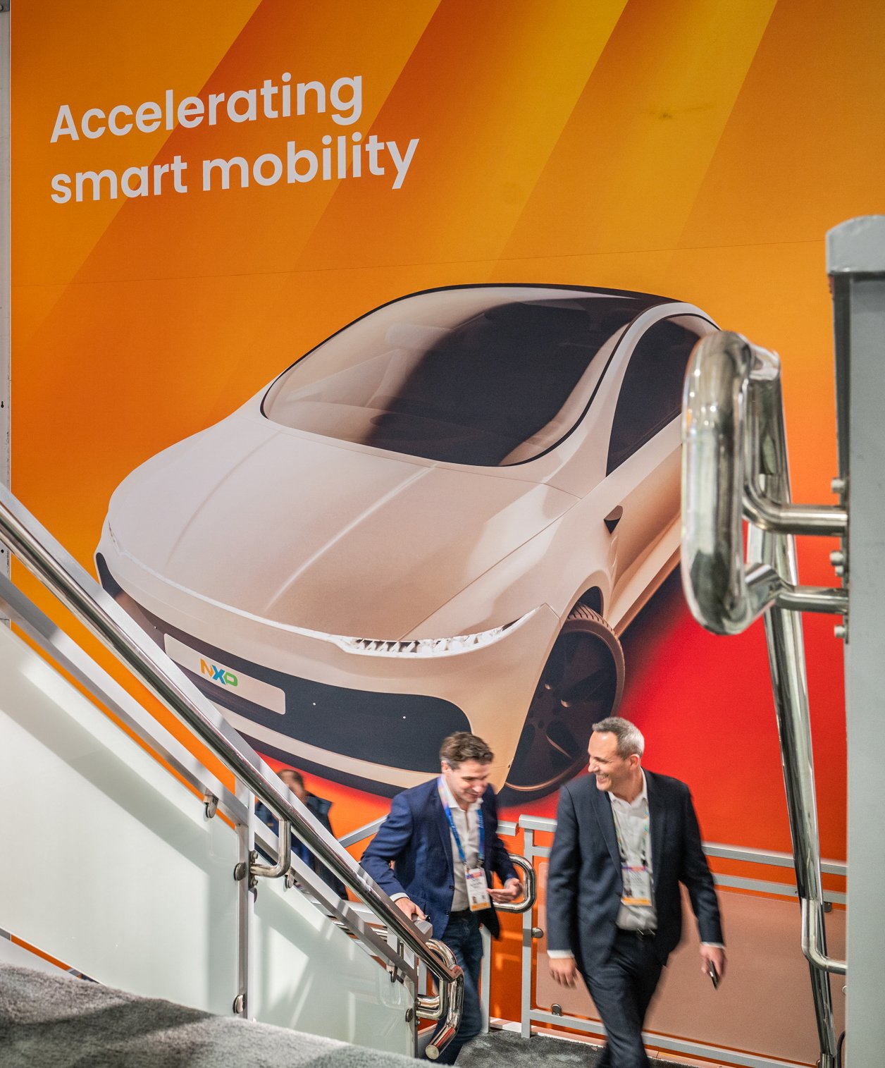 Two men walking down stairs in front of a large orange wall with a poster of a futuristic white electric car and the text 'Accelerating smart mobility'.