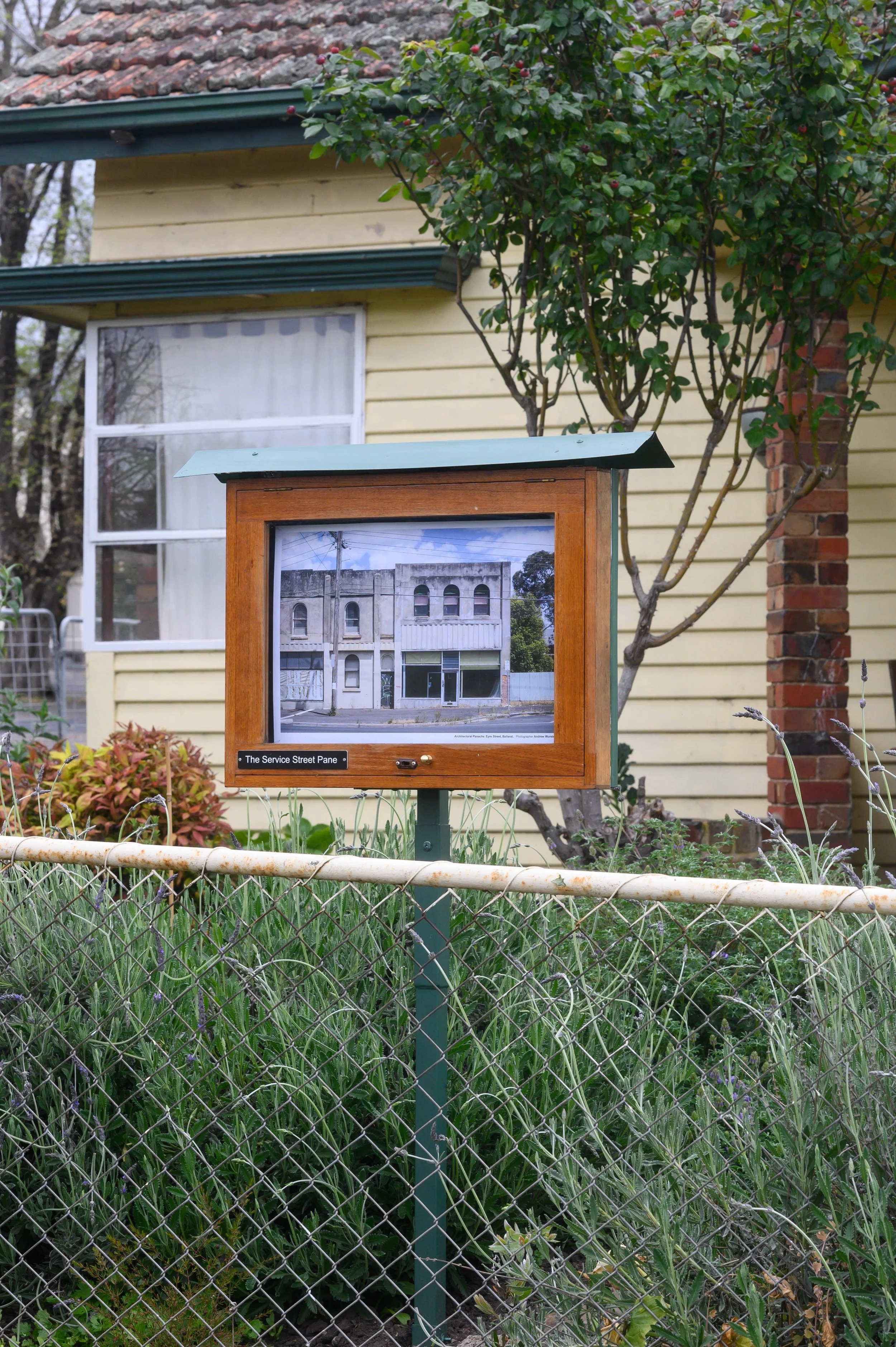 A street sign with a photo of a building, labeled 'The Service Street Pane,' is attached to a pole in a garden with green grass, plants, and a chain-link fence. A yellow house with a window, a tree, and a brick chimney are in the background.