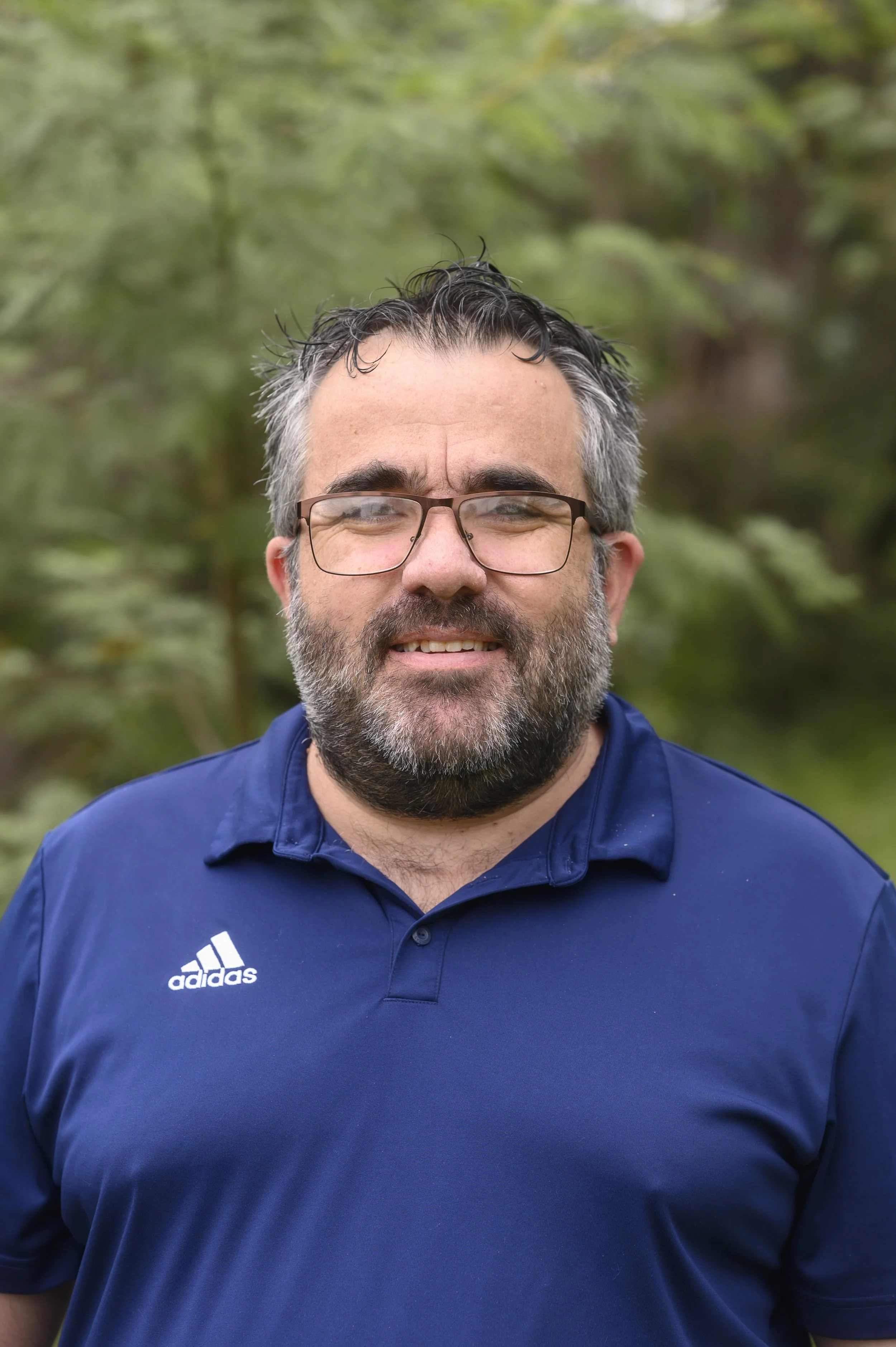 A middle-aged man with glasses, a beard, and gray hair, wearing a dark blue Adidas polo shirt, standing outdoors with green foliage in the background.