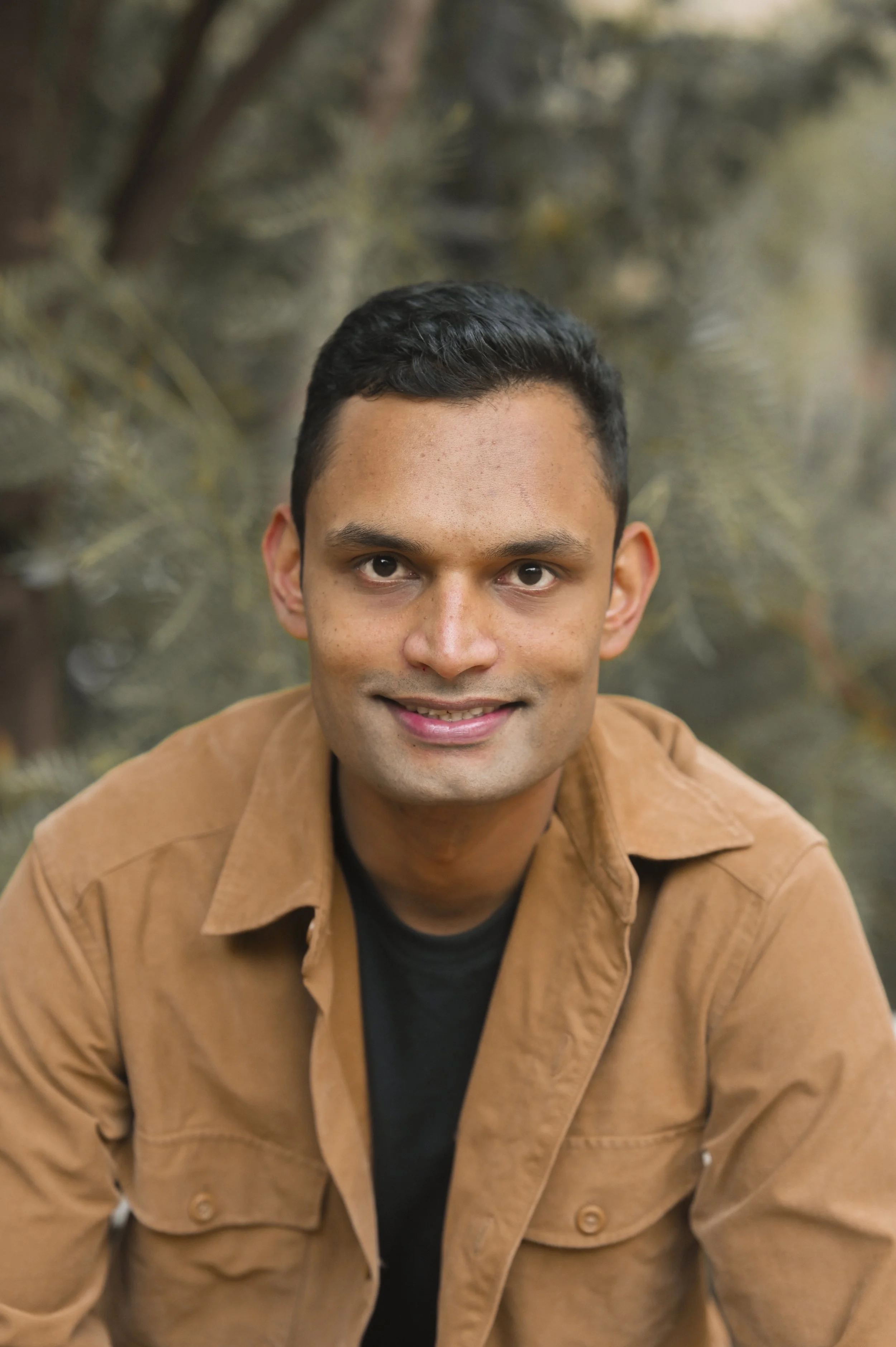 A smiling young man with short dark hair wearing a brown jacket over a black shirt, outdoors with blurred foliage in the background.