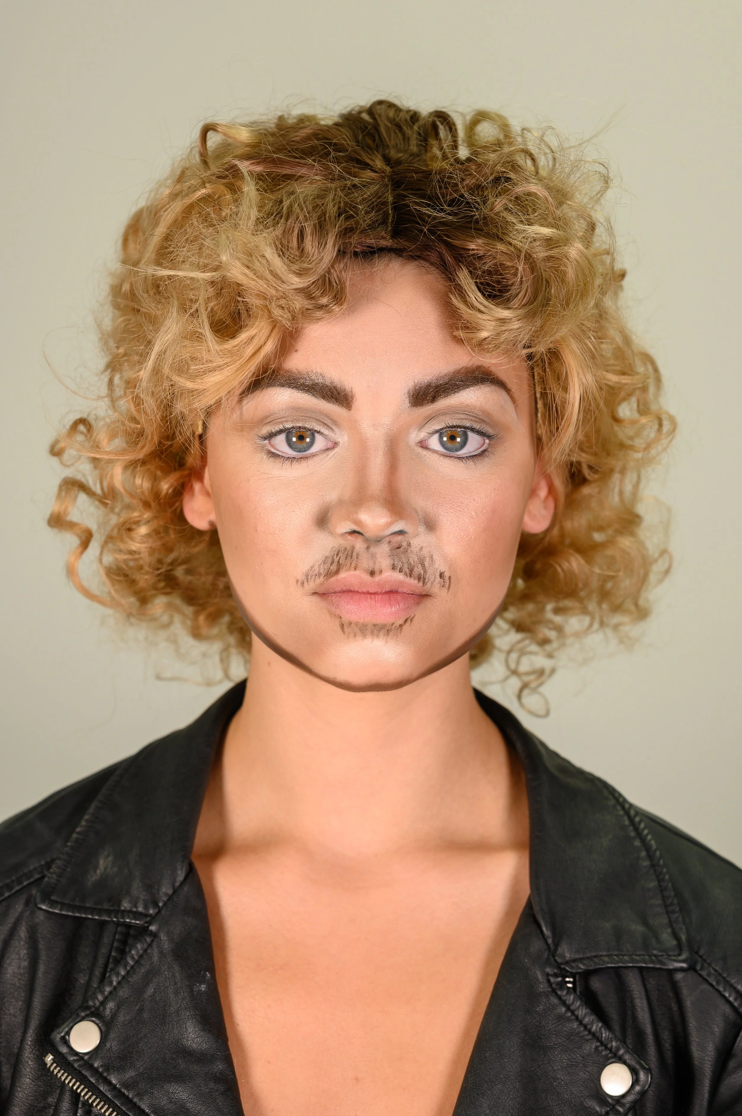 Person with curly blonde hair wearing makeup and a black leather jacket, with drawn-on facial hair, against a beige background.