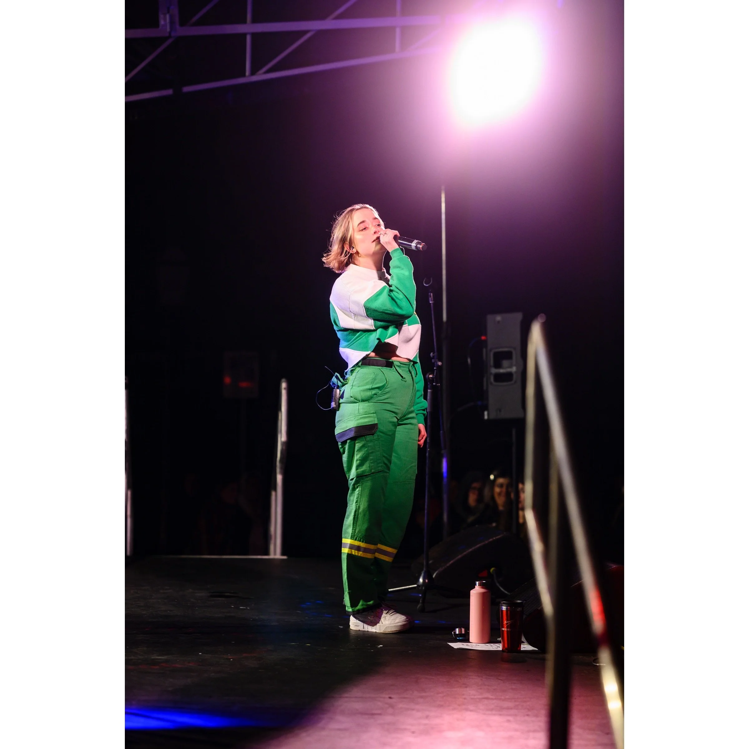 A woman singing into a microphone on stage with pink and purple stage lights, wearing a green and white track jacket and green cargo pants.