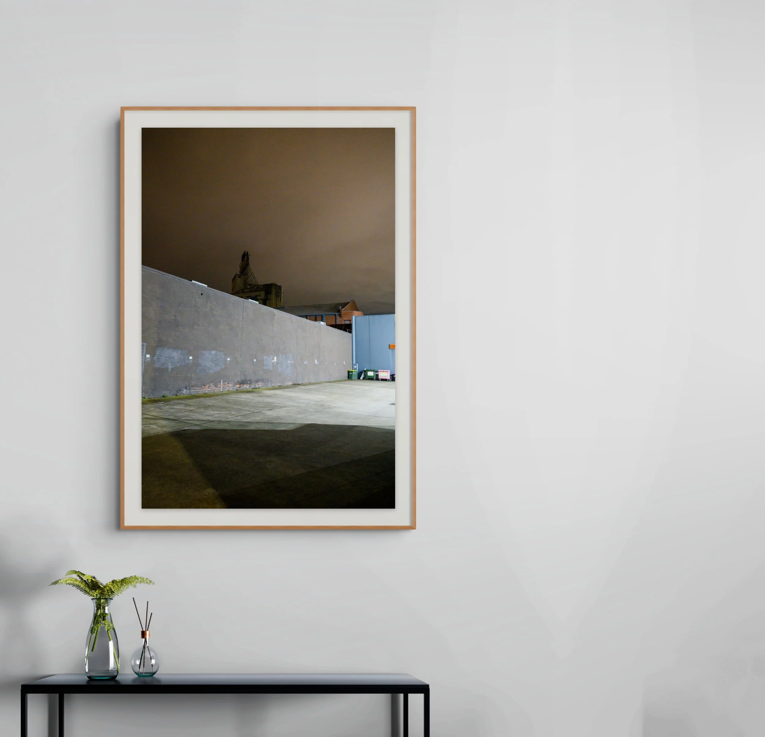 Minimalistic interior decor with a black table, two glass vases with green foliage, a framed photograph of a rooftop scene, and a small potted plant on the lower left corner of the image.