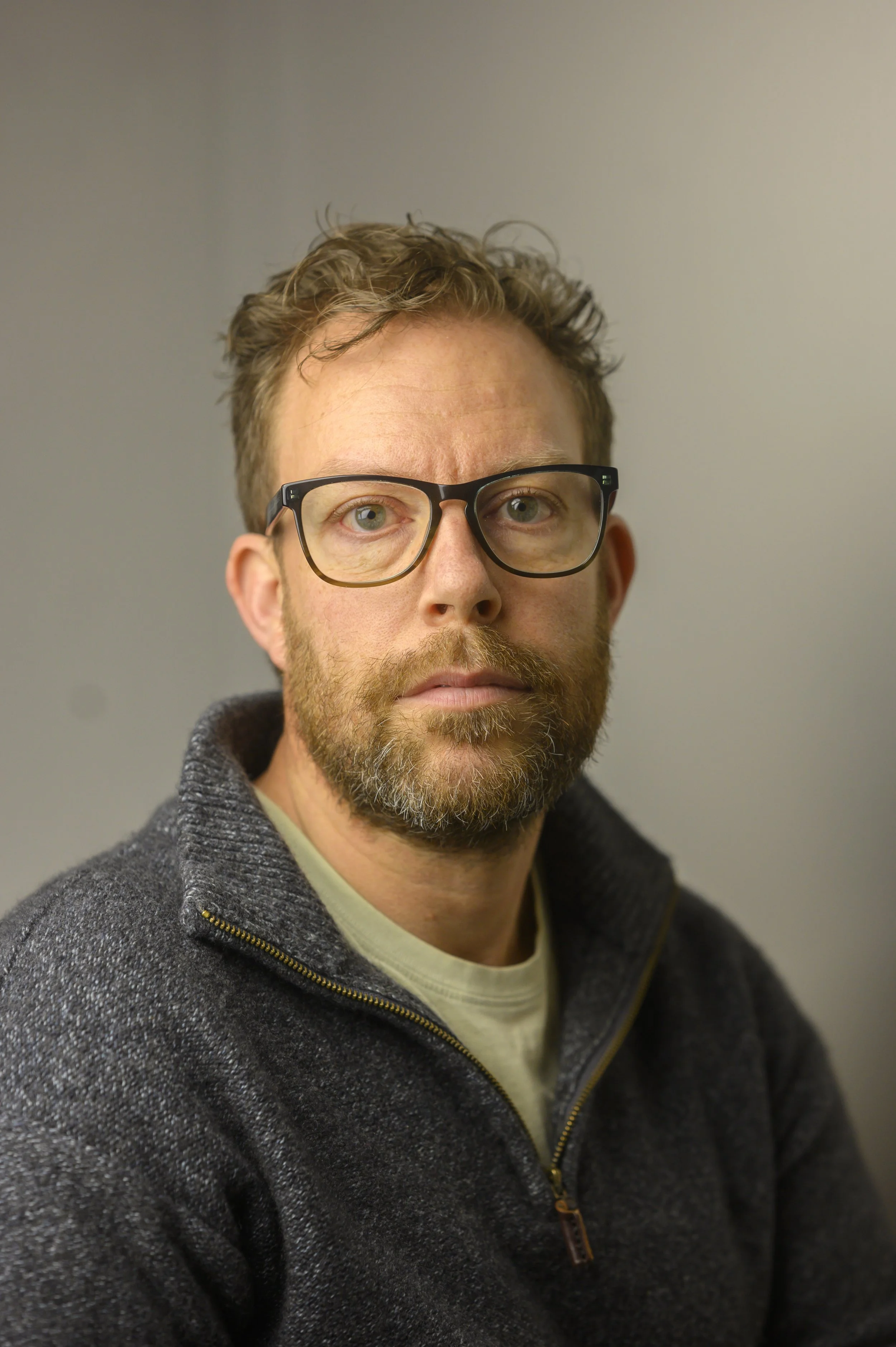 A man with glasses and a beard wearing a dark gray sweater, looking directly at the camera against a plain background.
