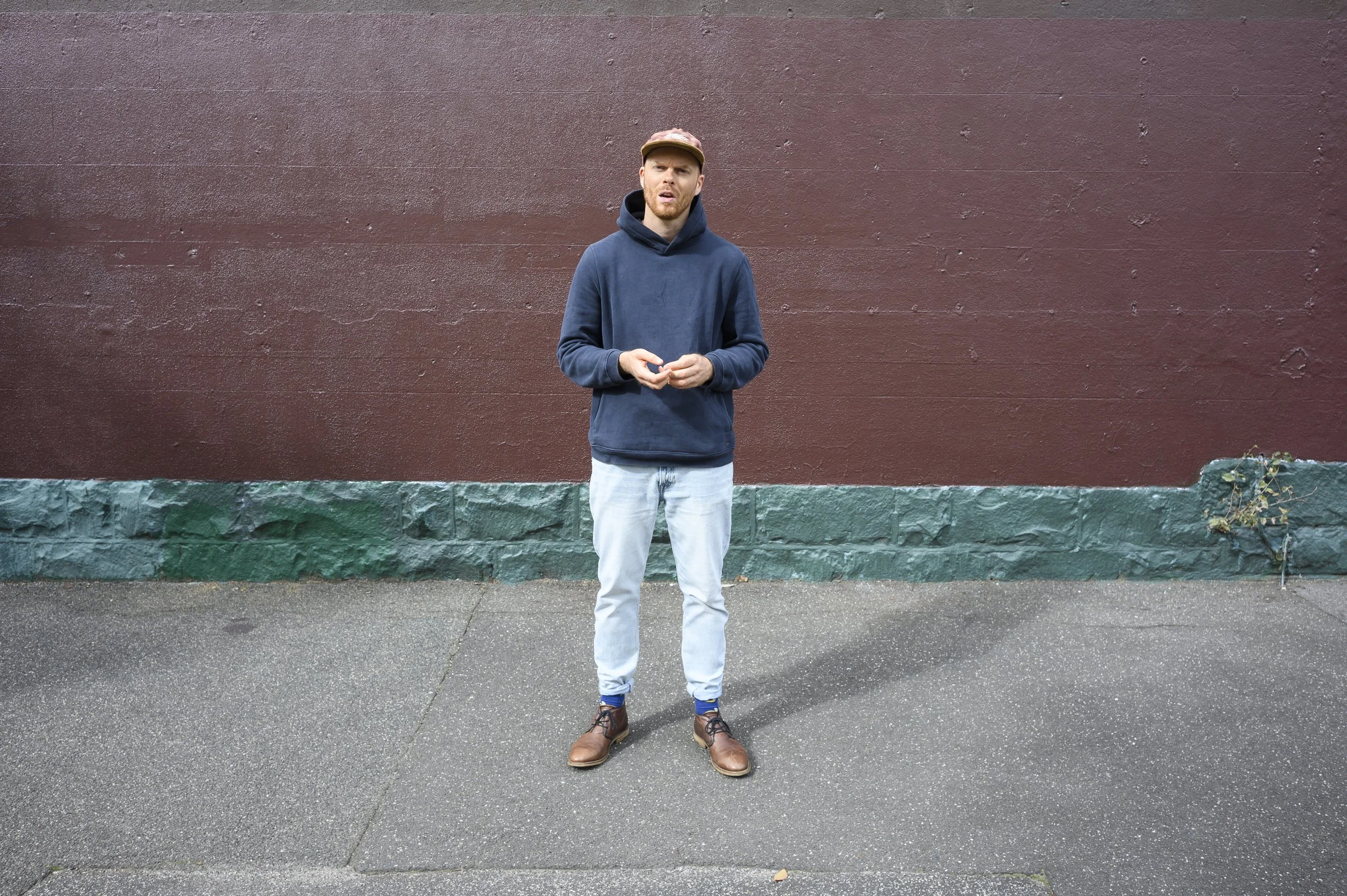 A man in a navy hoodie, light jeans, and brown boots standing on a sidewalk against a red wall with a green base, holding a small object in his hands and looking to the left.