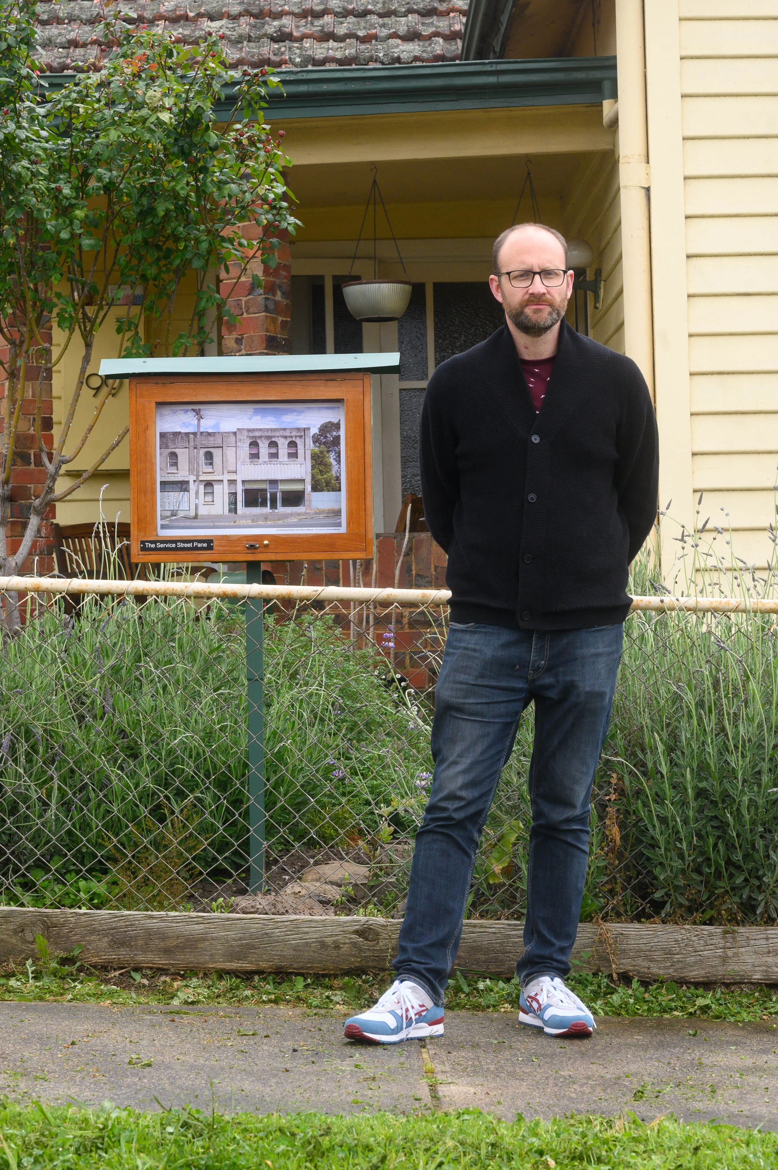 A man with glasses and a beard stands outdoors on a sidewalk, behind a small garden and next to an informational display board showing a building.