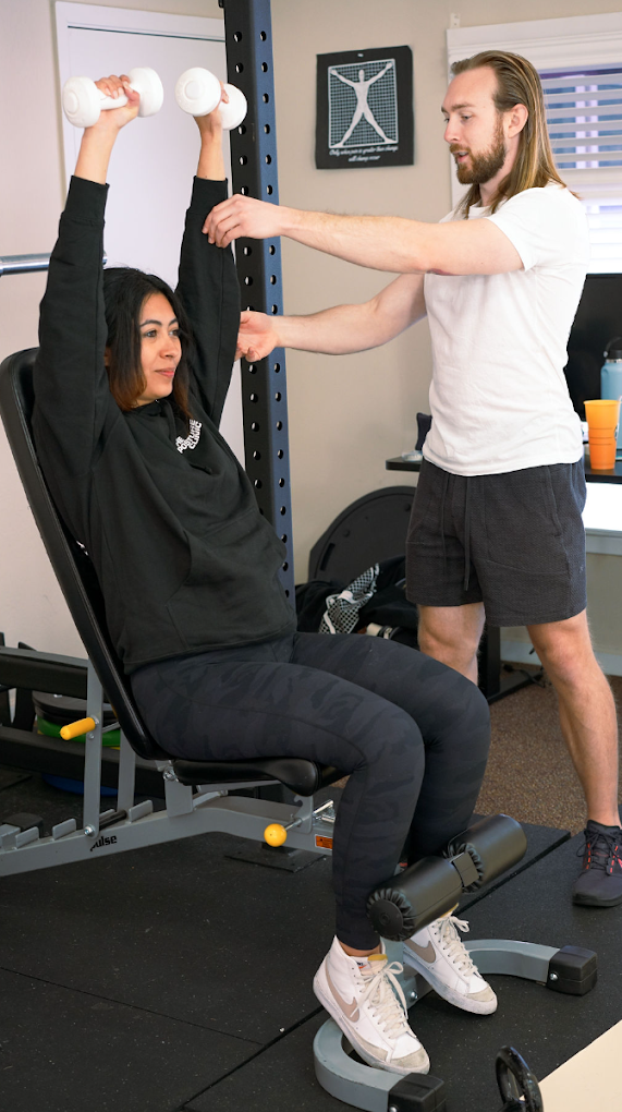 A woman sitting in a chair lifting dumbbells with her arms extended overhead while a male trainer stands beside her, providing guidance or assistance during a workout session in a gym.