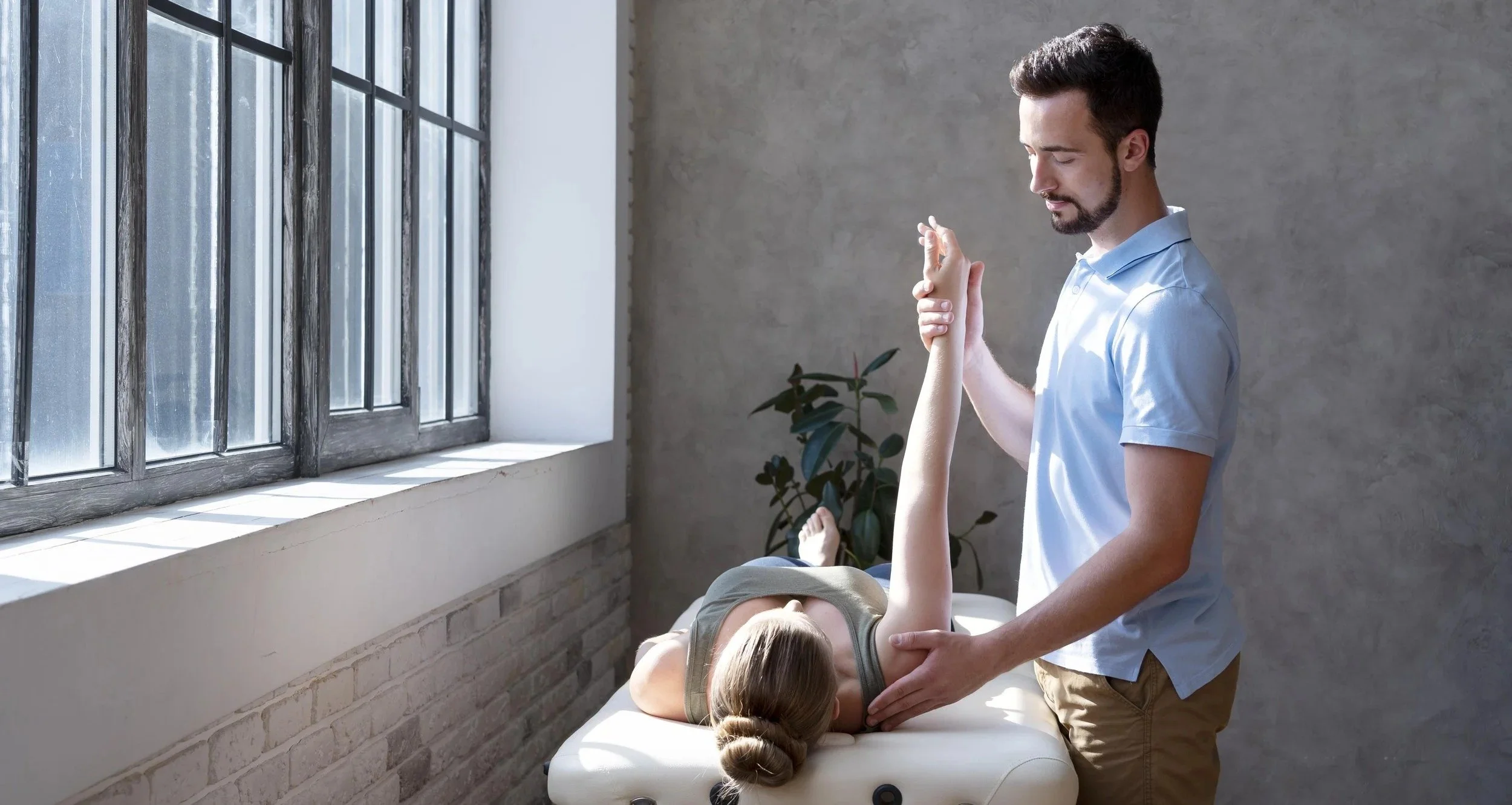 A chiropractor or physical therapist adjusting a woman's arm while she lies on a treatment table in a brightly lit room with large windows and a plant in the background.