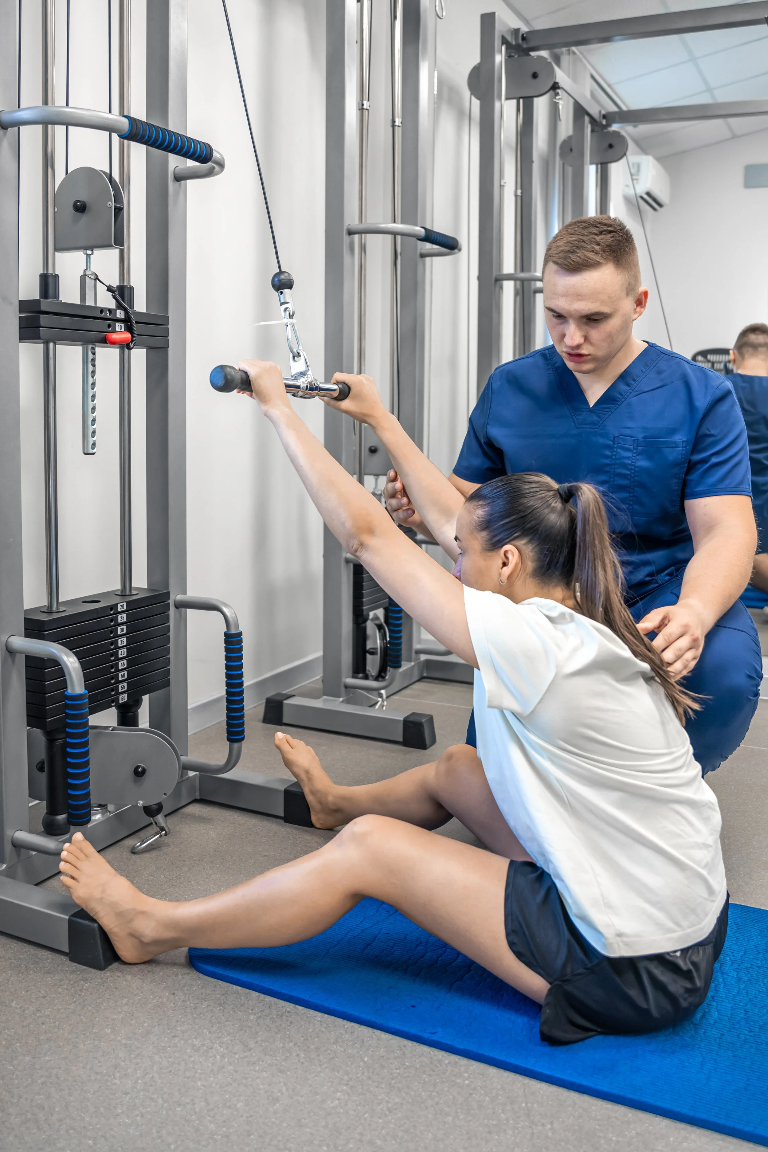 A young girl receives physical therapy from a male therapist at a gym, sitting on a blue mat while holding onto a cable machine for exercise.