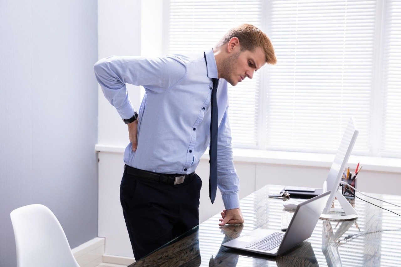 A man in business attire with a blue shirt and black tie standing in an office, leaning over a glass desk with a laptop, holding his lower back with one hand, appearing to experience back pain.
