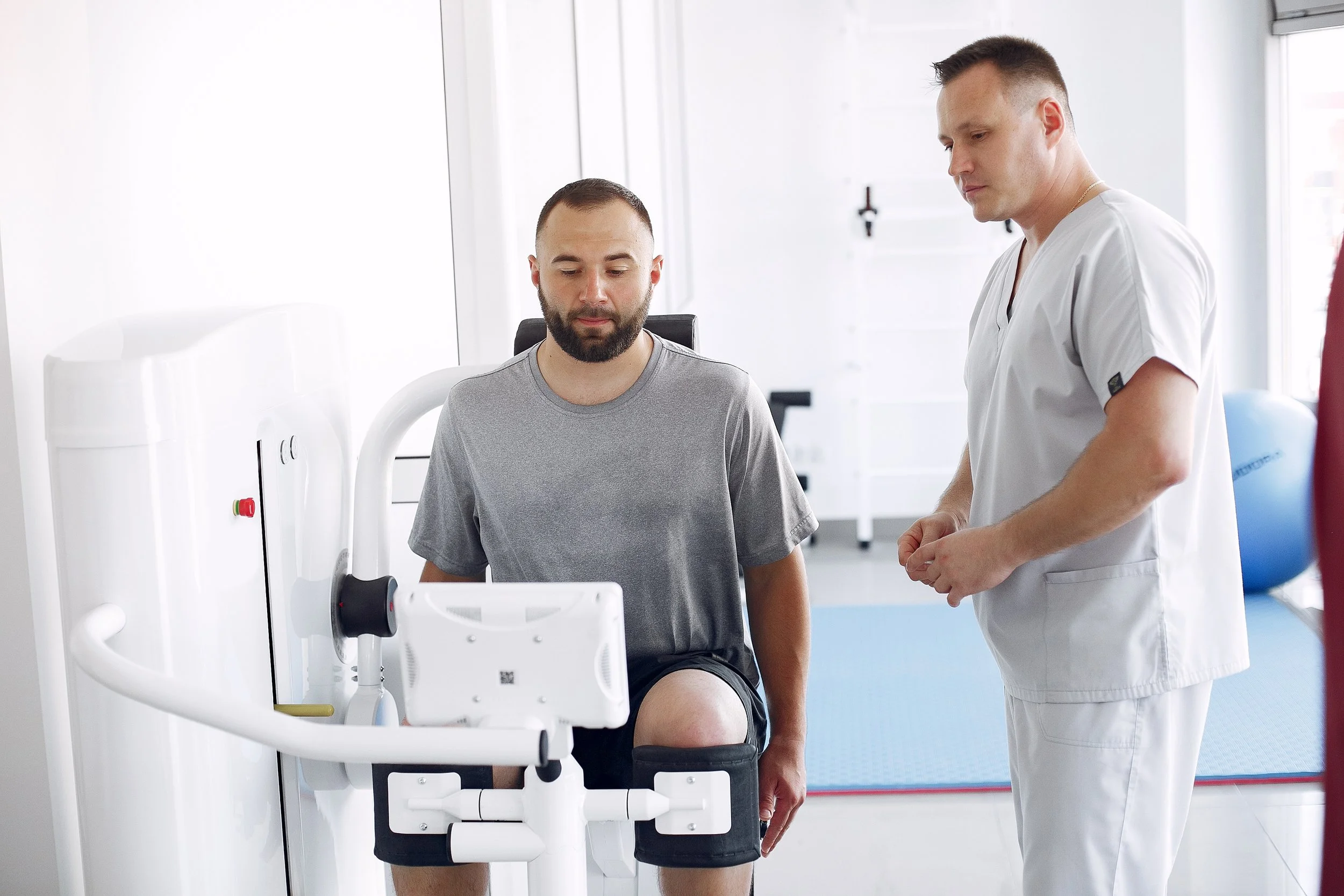 A man wearing a gray shirt sits on a chair with his knee placed on a knee machine, while a healthcare professional in white scrubs stands nearby, monitoring the process in a bright, medical environment.