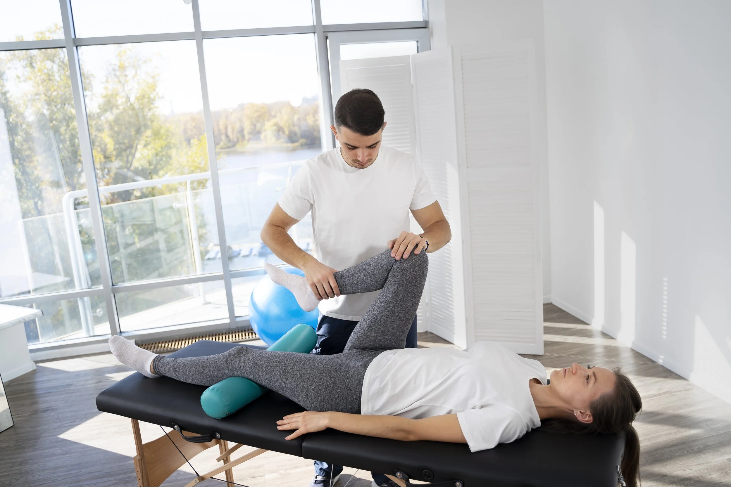 Male physical therapist assisting a female patient with leg exercises on a massage table in a bright room with large windows.
