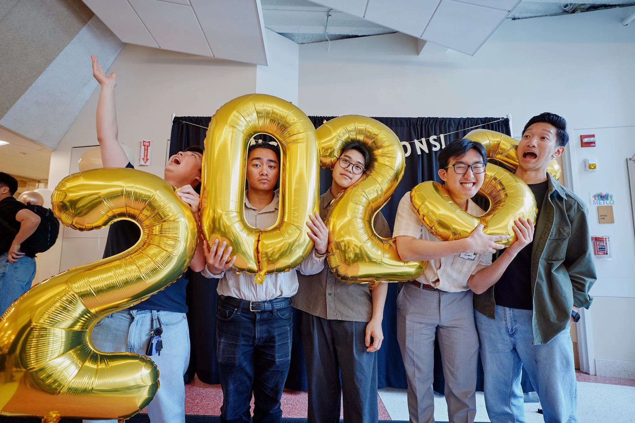Group of college students in Boston celebrating graduation with giant gold '2025' balloons, showing playful expressions at a church service community event.