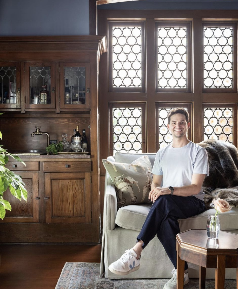 A smiling man in a white t-shirt and dark pants sitting on a beige couch inside a room with large windows featuring patterned wooden lattice. Behind him is a cozy throw blanket, and to his right is a small table with a flower in a glass vase. The room has wooden furniture, including a cabinet with bottles and glassware, and a potted plant is visible in the foreground.